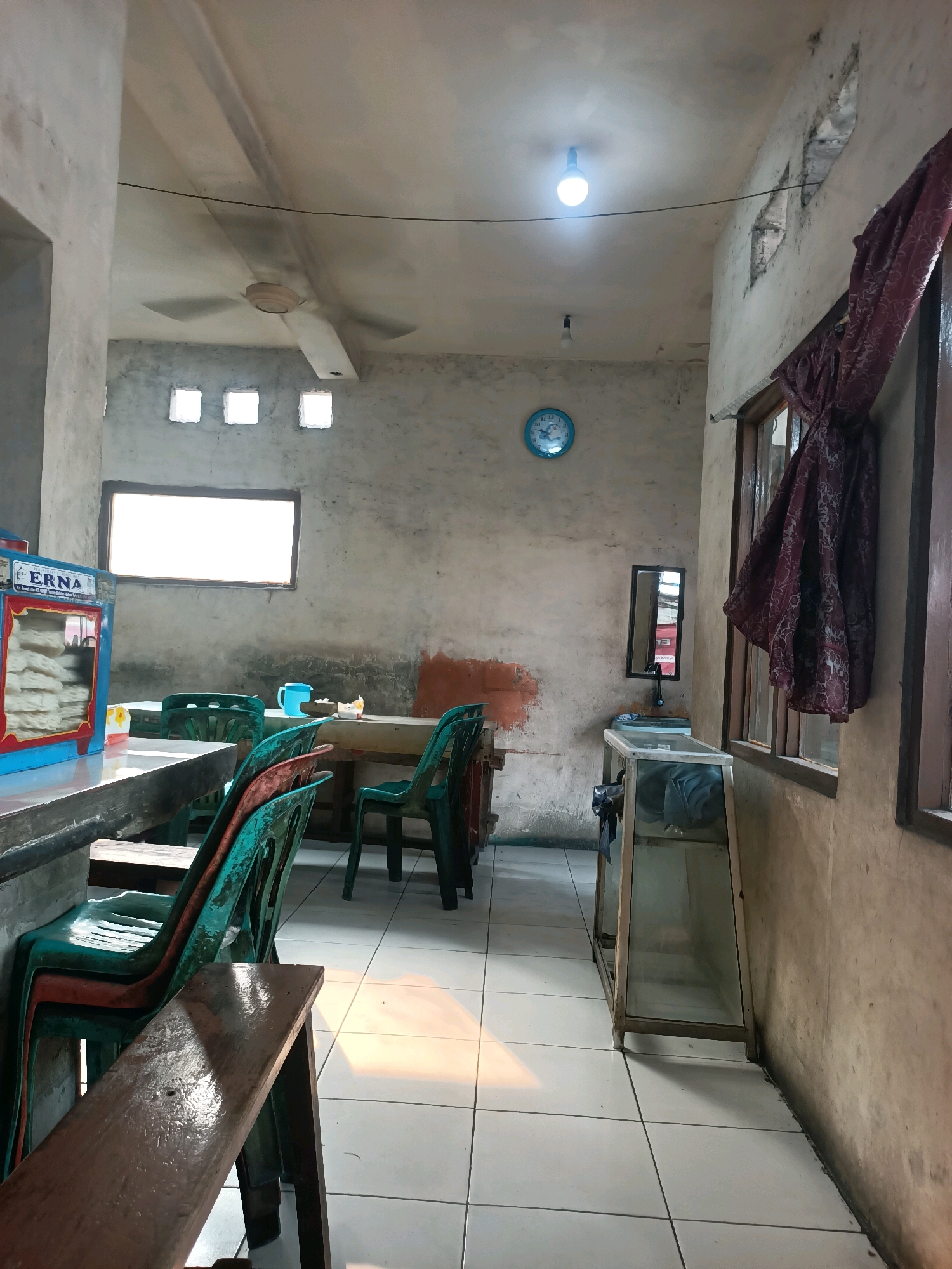 The image depicts the interior of a simple eatery in Gandasari, Indonesia. The walls are bare concrete, showing signs of age and minimal upkeep. A ceiling fan is visible overhead, along with a bare lightbulb illuminating the space. A round blue clock hangs on the wall.
In the foreground, a wooden bench is partially visible, with several stacked green plastic chairs to its right. Further into the room, there are tables with more green chairs. To the left, a food display case with the name "ERNA" is prominent, containing stacked flatbread or crackers.
A window with a white-blanked screen is set into the wall above one of the tables. On the right side of the image, a tall, narrow window with wooden frames is partially covered by a dark, patterned cloth. Below this window, a glass-fronted display cabinet stands on the floor. A small mirror is mounted on the wall above a simple sink area. The floor is tiled with white square tiles. The overall impression is of a functional, unadorned dining space.