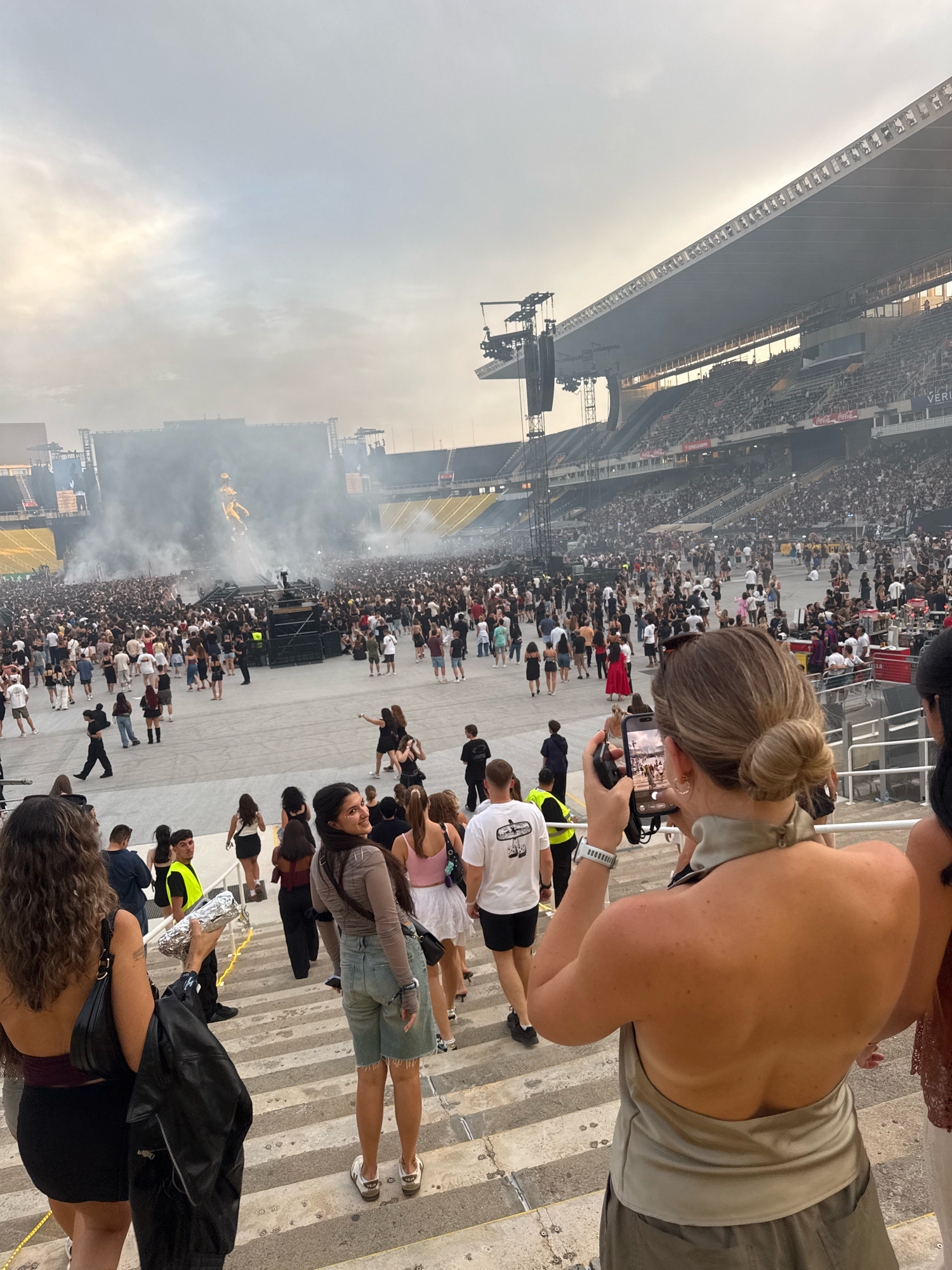 The image captures a wide view of a large crowd gathered in a stadium, presumably for an outdoor concert or event, under a hazy sky. In the foreground, several individuals are visible on stadium steps, with some walking up and others standing. To the right, a woman is taking a photograph with her phone, her back to the camera, showcasing an open-back, light-colored halter top. Further to the left, another woman, facing the camera and smiling, is wearing a brown long-sleeved top and denim shorts. She is holding a silver-wrapped object in her right hand. In the background, a massive crowd stretches towards a stage set up in the center of the stadium. Smoke or fog emanates from the stage area, obscuring parts of the scene. Multiple large screens and lighting rigs are visible above the stage. Stadium seating, filled with spectators, surrounds the performance area. The overall atmosphere suggests a lively event with a significant turnout. Although the prompt asks to include the city of Barcelona, Spain, there are no visual cues in the image to confirm this specific location.