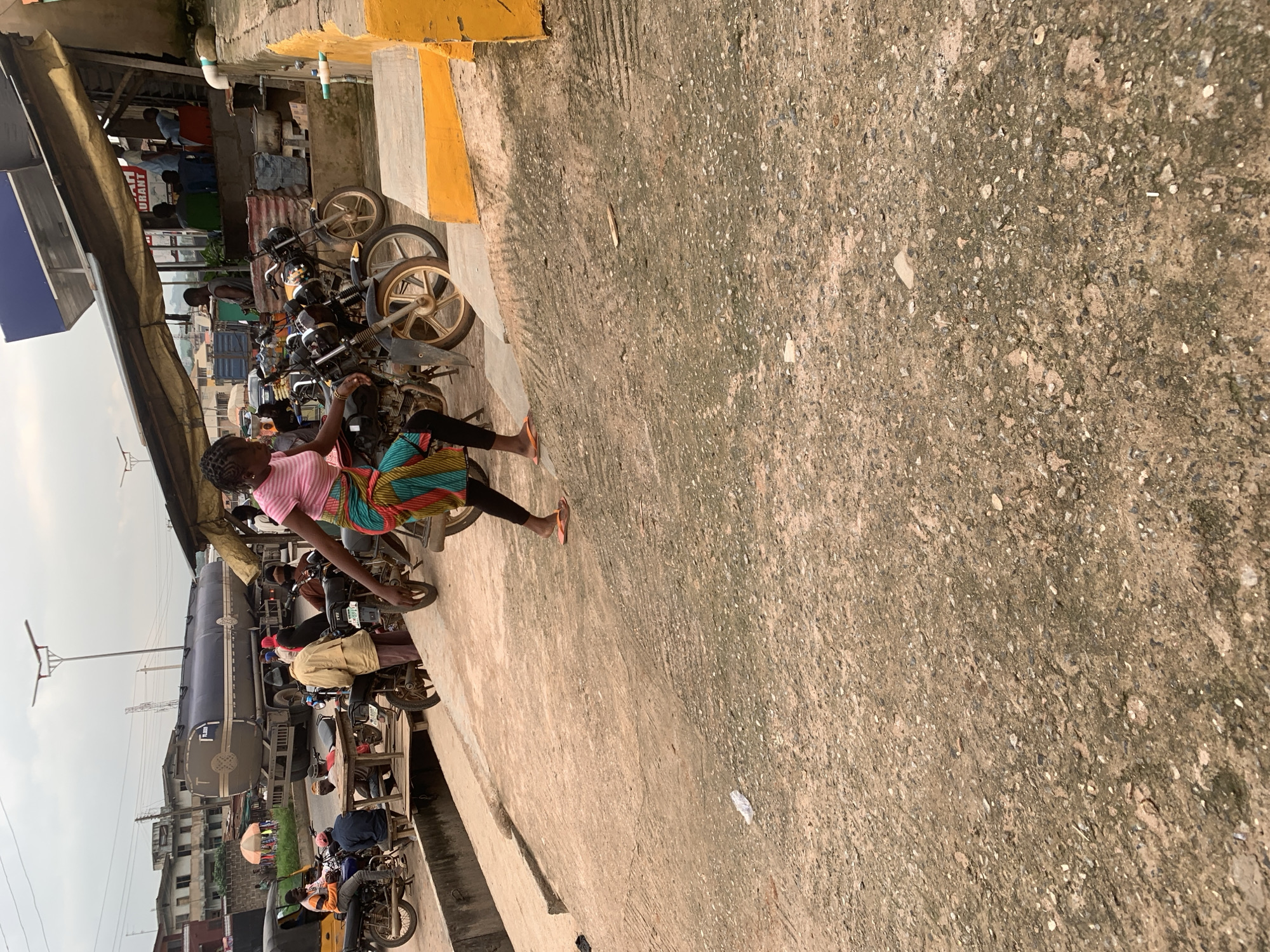 The image captures a street scene in Shagamu, Nigeria, focusing on a woman in a pink top and a colorful patterned skirt walking past parked motorcycles. She is wearing orange flip-flops and appears to be moving downhill. In the background, several motorcycles are visible, some with riders. Further back, a large tanker truck is parked on the road, and buildings line the street. The environment appears to be a commercial area, possibly a market or a roadside gathering spot, with a covered structure and seating in the distance. The ground is paved with concrete, showing signs of wear and tear. The overall atmosphere is that of a busy, everyday urban environment.