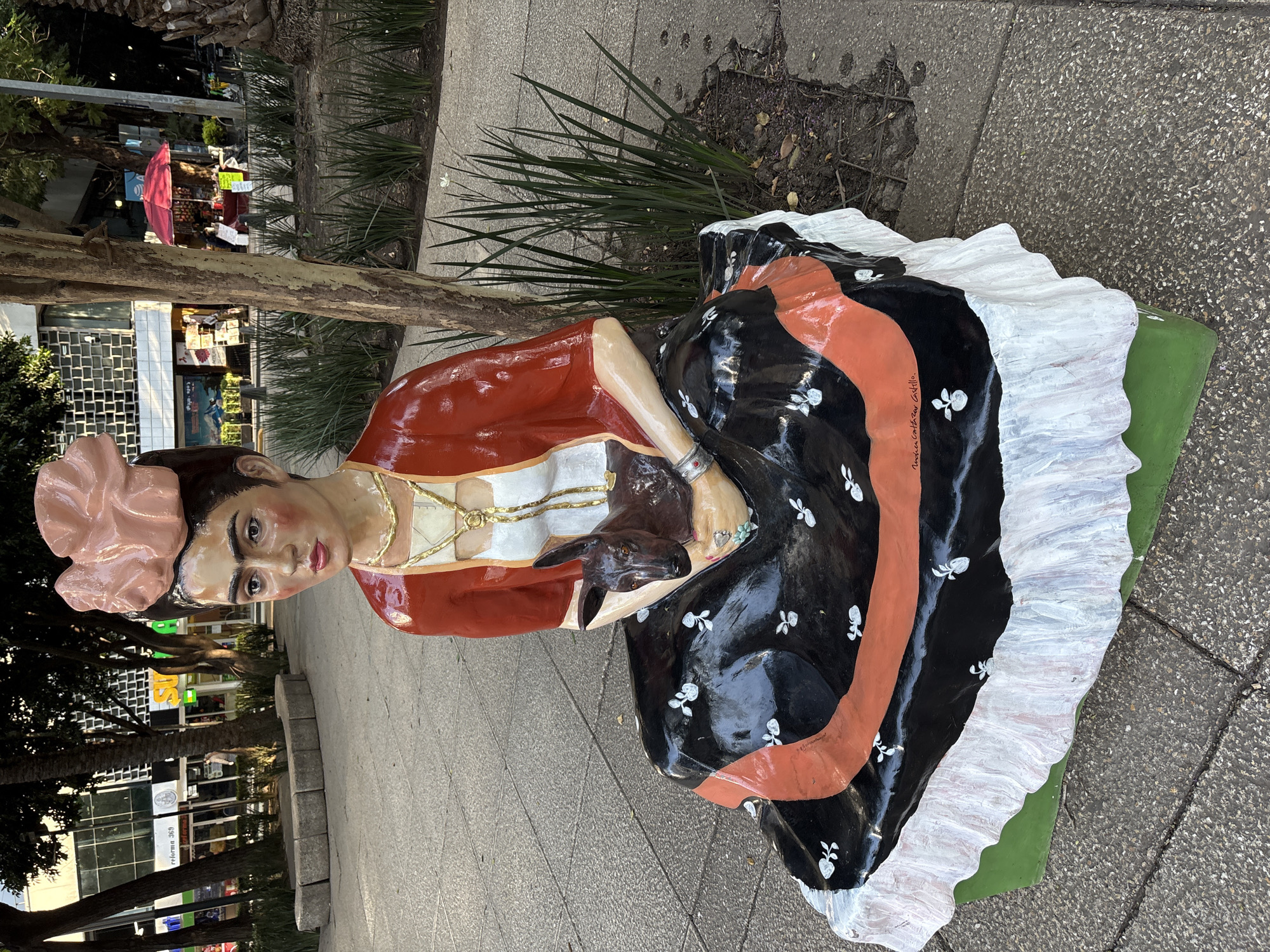 A life-sized statue of Mexican artist Frida Kahlo is seated on a bench in a public outdoor area in Mexico City, Mexico. The statue depicts Kahlo in her iconic style, wearing a traditional dress and holding a small animal, possibly a deer or dog, in her arms. She is adorned with a large floral headdress and elaborate necklaces, and her unibrow and distinct features are prominent. The statue is positioned in front of a tree with lush green foliage and on a paved walkway with stone borders. In the background, there are buildings, street vendors with umbrellas, and more greenery, suggesting an urban park or plaza setting. The statue's base is painted bright green with a white ruffled skirt detail. The overall scene captures a piece of public art integrated into the city's landscape.