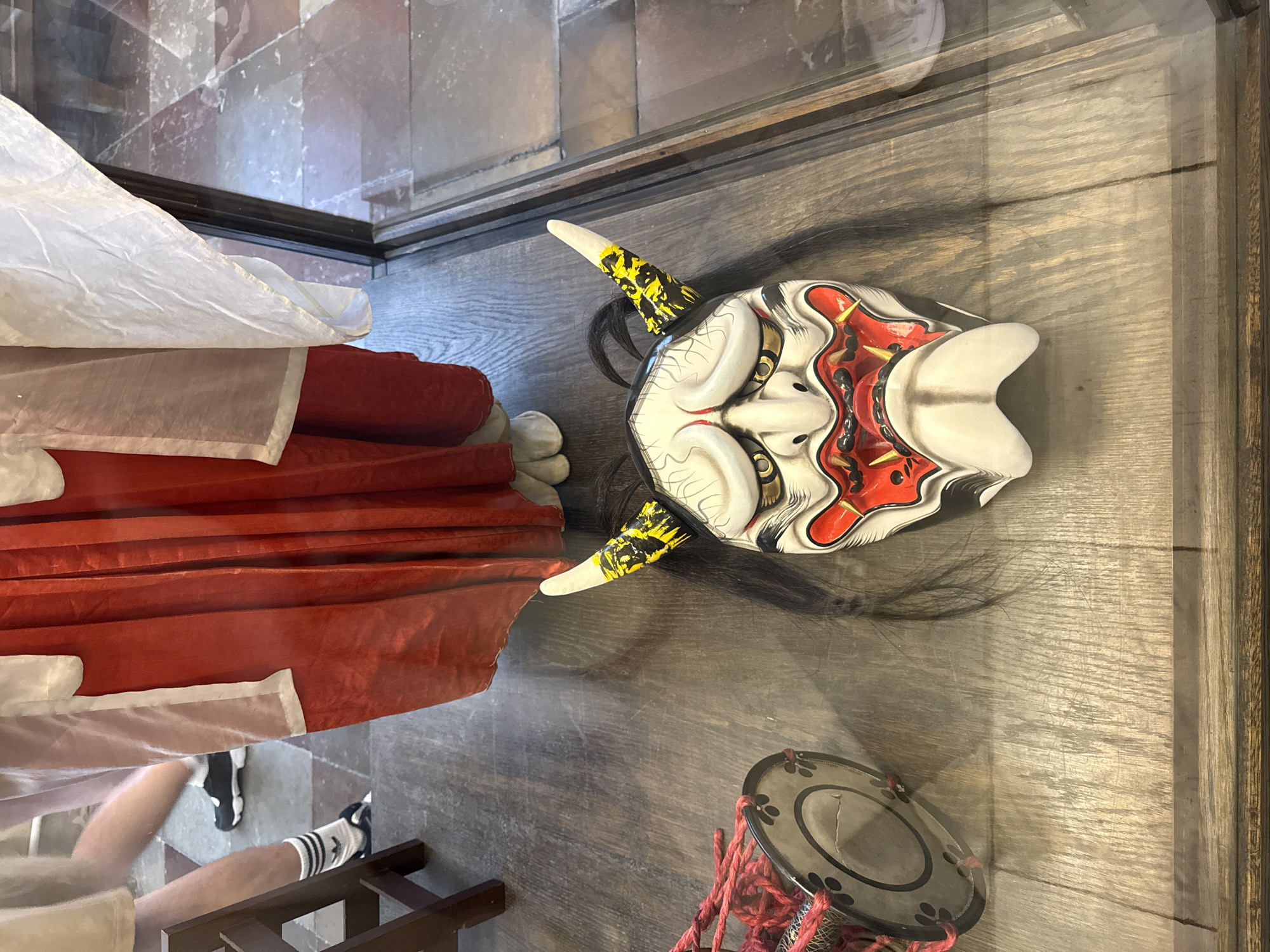 An elaborately decorated Japanese Oni mask, with black hair and yellow and black striped horns, rests on a wooden floor next to a small drum with red rope. The mask is viewed through a glass enclosure, possibly a display case within a museum. To the left of the mask and drum, the lower legs and feet of two individuals are visible. One person is wearing black sneakers with white socks, and the other is wearing white athletic shoes with black accents. Behind the mask and to the right, a portion of a mannequin or figure dressed in a red and white garment is visible. The floor appears to be wood planks with a textured, worn surface. The background shows a tiled floor and part of a glass display case with a dark wooden frame. The context of Saint Petersburg, Russia is not evident from the visual information in the image.