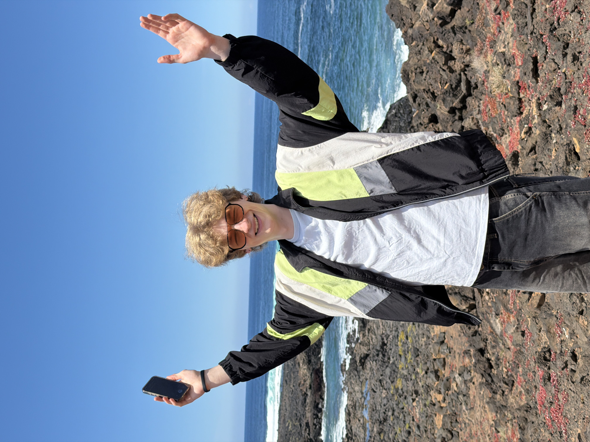 A young, blond-haired male is standing on a rocky outcrop overlooking the ocean, possibly along the coast of Tinajo, Spain. He is wearing sunglasses, a white t-shirt, a multi-colored windbreaker (black, white, grey and lime green), and jeans. His arms are outstretched, one holding a phone. The background includes the ocean and a clear blue sky. The ground near him is rocky and covered with some vegetation.
