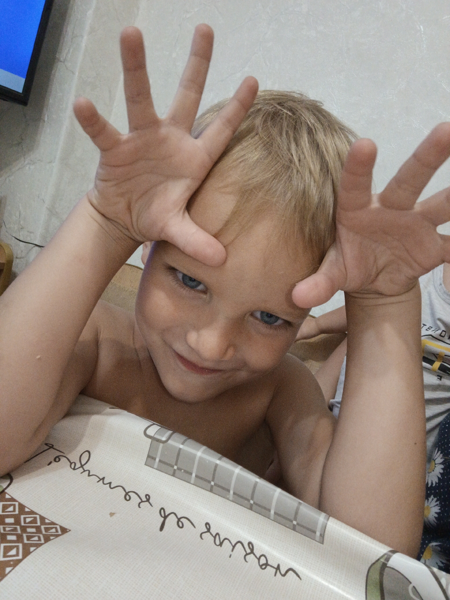 This is a close-up, medium shot of a young boy with blonde hair and blue eyes. He is shirtless and has his hands raised in front of his face, fingers splayed, in a playful gesture. His mouth is slightly open in a smile, and he is looking directly at the camera. The boy is leaning over a surface with a patterned tablecloth. The background appears to be a wall with a textured surface, and a portion of a television screen is visible in the upper left corner, showing a bright blue image.
The activity appears to be a posed photograph, with the boy making a funny face for the camera. The lighting suggests it is daytime, possibly indoors due to the presence of the television and indoor setting implied by the tablecloth. There are no obvious cues for weather or specific time of day beyond it being light.
The tablecloth has a repeating pattern of geometric shapes and cursive writing. While the writing is not entirely clear, some of the letters appear to be Cyrillic, consistent with the location context of Medvedyovka, Russia.