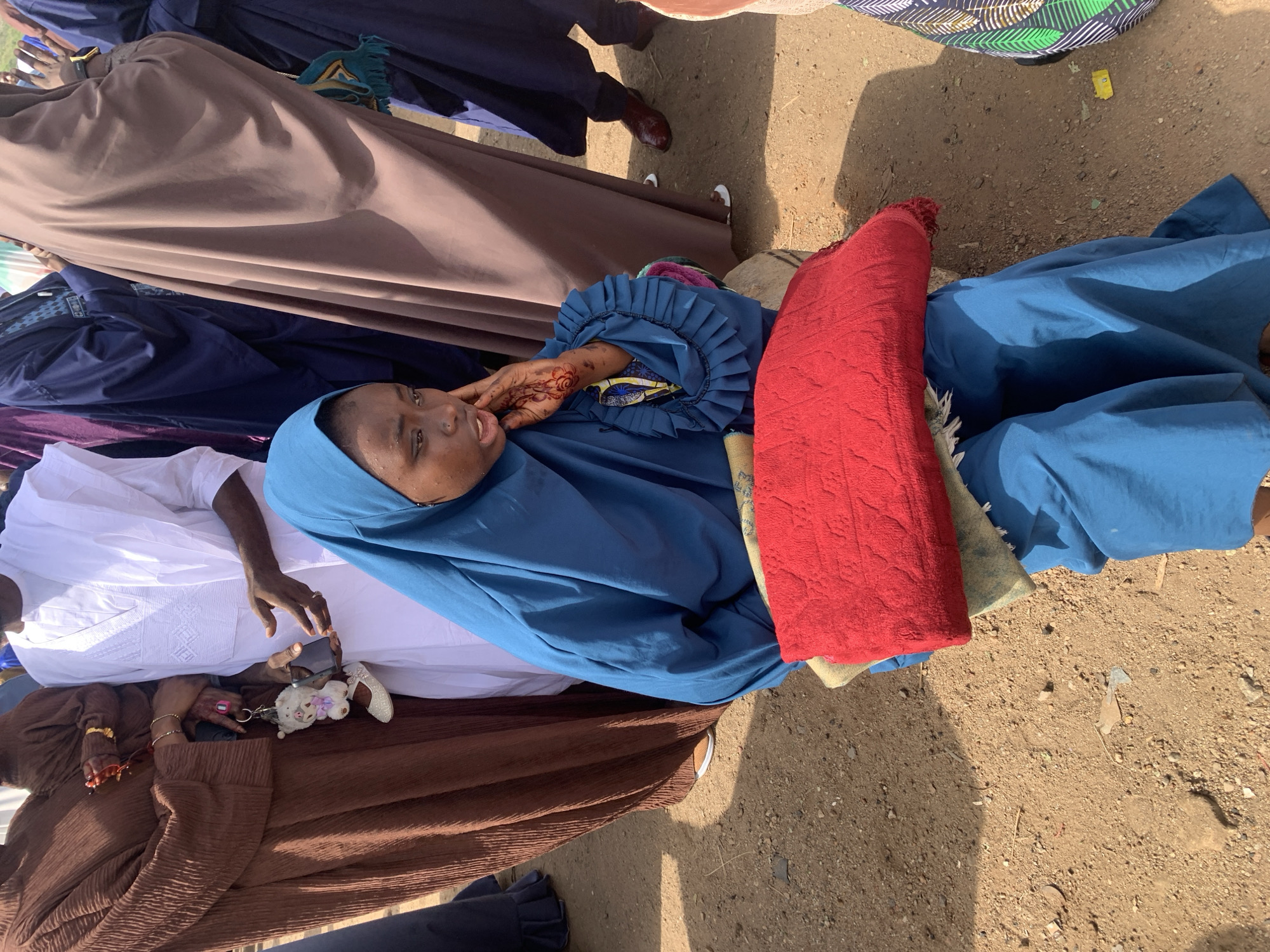 The image shows a gathering of people in what appears to be an outdoor location in Naraguta Hausawa, Nigeria. Predominantly women are visible, dressed in traditional Islamic attire, including headscarves (hijabs) and gowns in varying colors such as blue, brown, and white. One woman is lying down, resting her head on a red fabric. Her hand has henna designs on it. Other people are standing around her. The ground is dirt or gravel. Some accessories such as keychains are visible in people’s hands.