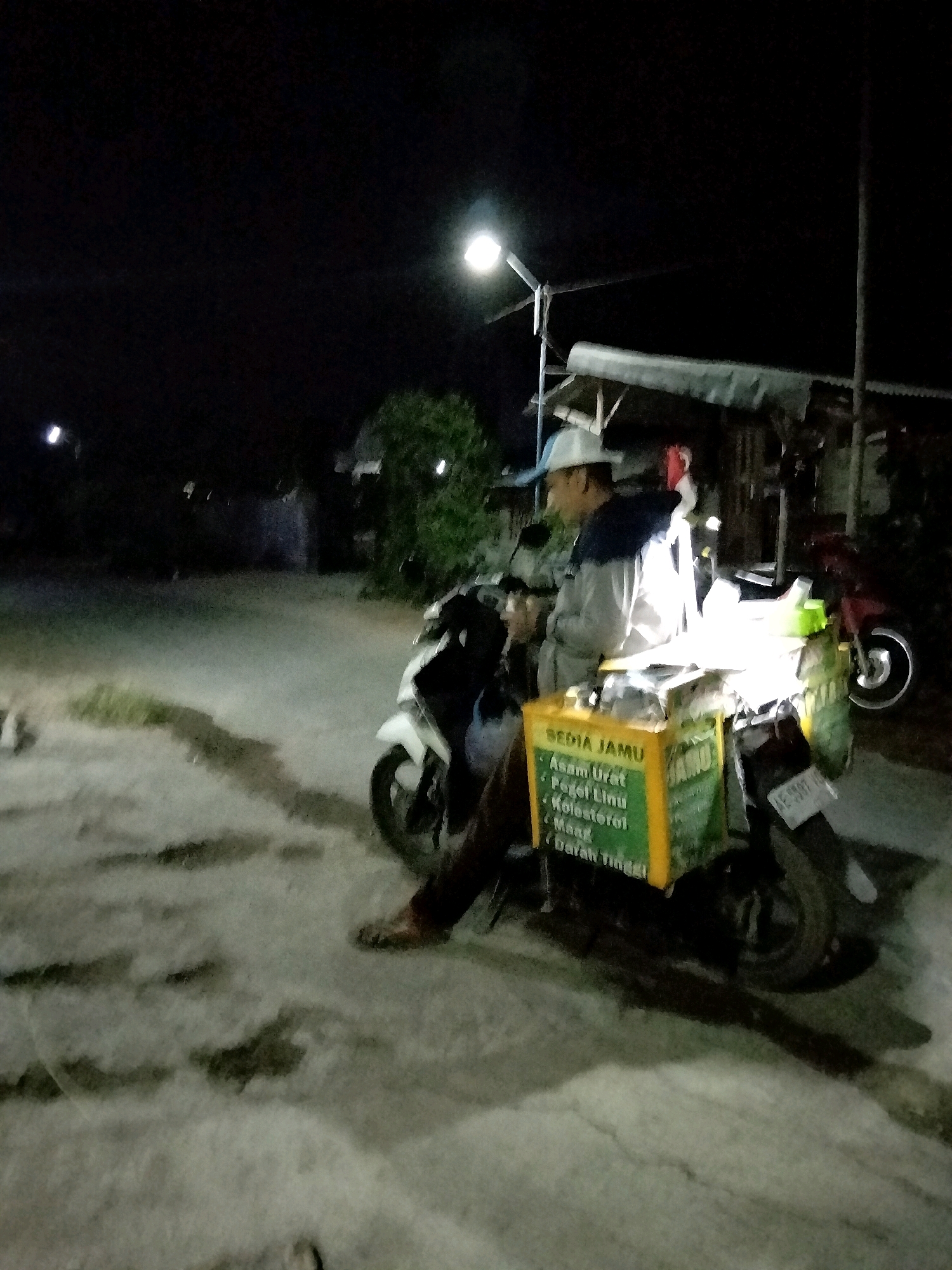 At night in Mrican, Indonesia, a man wearing a white cap and a blue and gray jacket sits on a motorcycle. The motorcycle has a green and yellow box attached to its back, which appears to be a vendor's stall. The box has text written on it that includes "SEDIA JAMU" and a list of ailments such as "Asam Urat," "Pegel Linu," "Kolesterol," "Maag," and "Darah Tinggi." The man is looking to his left, and the motorcycle is parked on a paved surface that is illuminated by a nearby streetlamp and the lights from the stall. In the background, there are some trees and structures, suggesting a roadside setting.