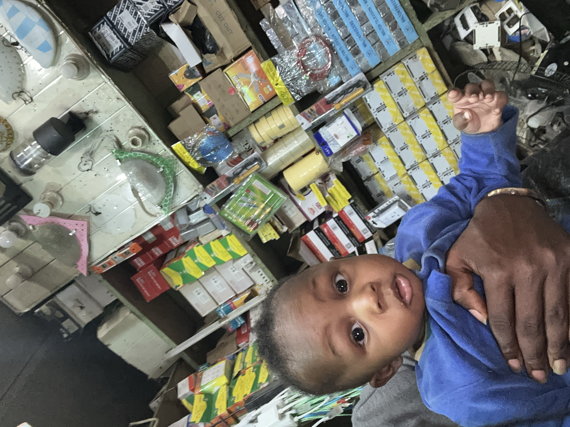 The image depicts a young baby held by an adult within what appears to be a cluttered retail store located in Narayi, Nigeria. The baby, with dark skin and short, dark hair, wears a blue long-sleeved garment and looks directly at the camera with wide, open eyes and a slightly parted mouth. A dark-skinned adult hand, adorned with a gold-colored bracelet, supports the baby's torso.
The background consists of multiple densely packed shelves filled with various merchandise. On the left, bundles of green, white, and blue wires are visible. Further back, numerous cardboard boxes in yellow, green, and red hues are stacked. Rolls of yellow and clear adhesive tape are present on a mid-level shelf. Various packaged items, including what appear to be soldering irons and electrical components (some labeled "WALTON TELEVISION," "WALTON FREEZERS," and "WALTON GAS COOKER"), are arranged throughout the shelves. An upper shelf displays several light fixtures or components, including a black cylindrical item, white dome-shaped objects, and a pink, patterned decorative item. The overall environment suggests a hardware, electronics, or general merchandise shop.