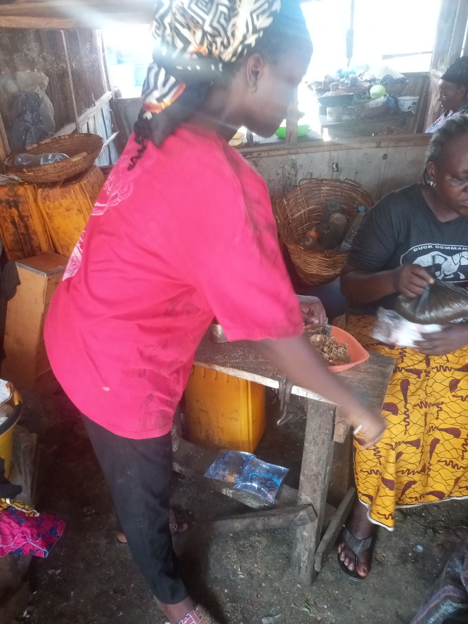 A scene in Sabon Tasha, Nigeria, shows two women preparing food. One woman wears a patterned headscarf, a red shirt, and black pants. She stands at a rustic wooden table, using a utensil to serve food from an orange bowl. The other woman sits with a yellow and brown patterned skirt and a dark T-shirt with a white graphic. She holds a plastic bag. Behind them are baskets and yellow containers. The floor appears to be dirt.