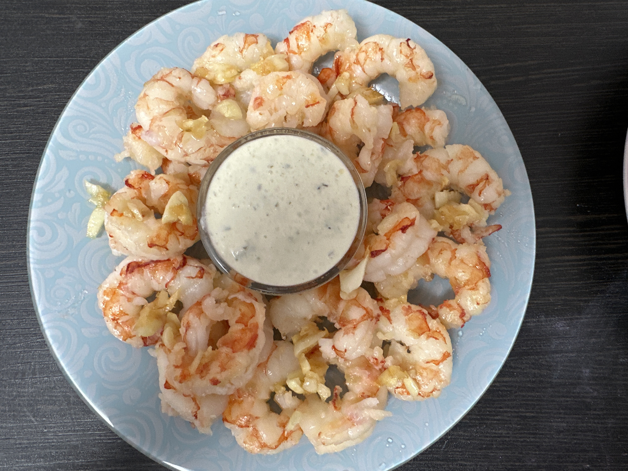 A close-up, overhead shot showcases a light blue, circular plate with a swirled pattern, filled with cooked shrimp. A small glass bowl of creamy white sauce with green specks sits in the center of the plate, surrounded by the shrimp, which are a pale pink and white with hints of orange. Thin slices of garlic are scattered amongst the shrimp. The plate rests on a dark, wooden surface. The image does not contain any elements that would indicate a specific location in Chaikovsky, Russia.