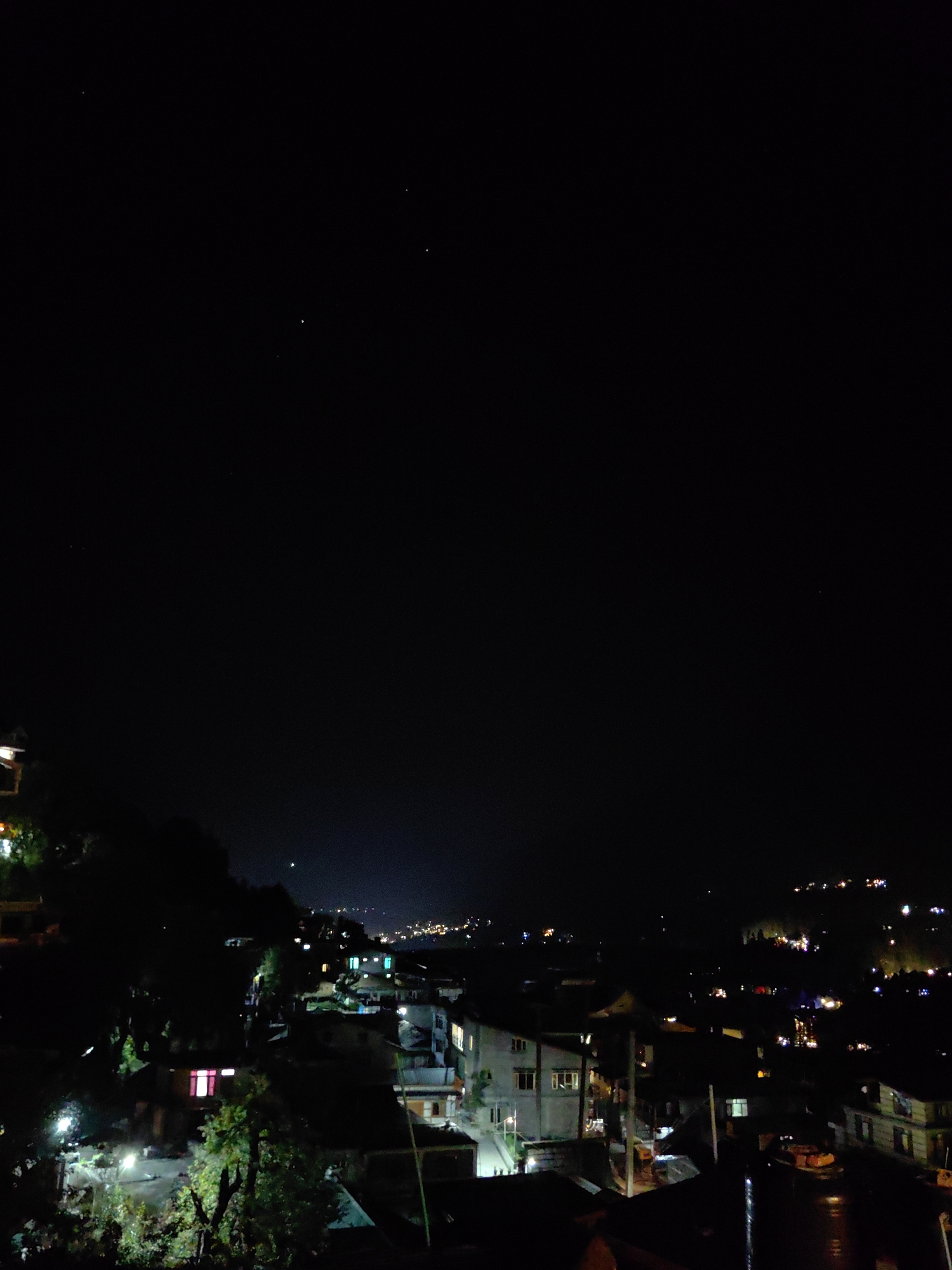 This image depicts a nighttime view of a hillside town, likely Vashisht, India, characterized by its numerous houses with illuminated windows. The foreground features darker, silhouetted trees and structures, with some buildings showing distinct lights, such as a pink-hued window. The midground reveals a cluster of residential buildings, some with visible lighting and architectural details. The background shows a sprawling cityscape of lights extending up a darkened hillside, suggesting a developed settlement. A few faint stars are visible in the inky black sky above. The overall impression is of a vibrant community nestled in a mountainous landscape after dark.