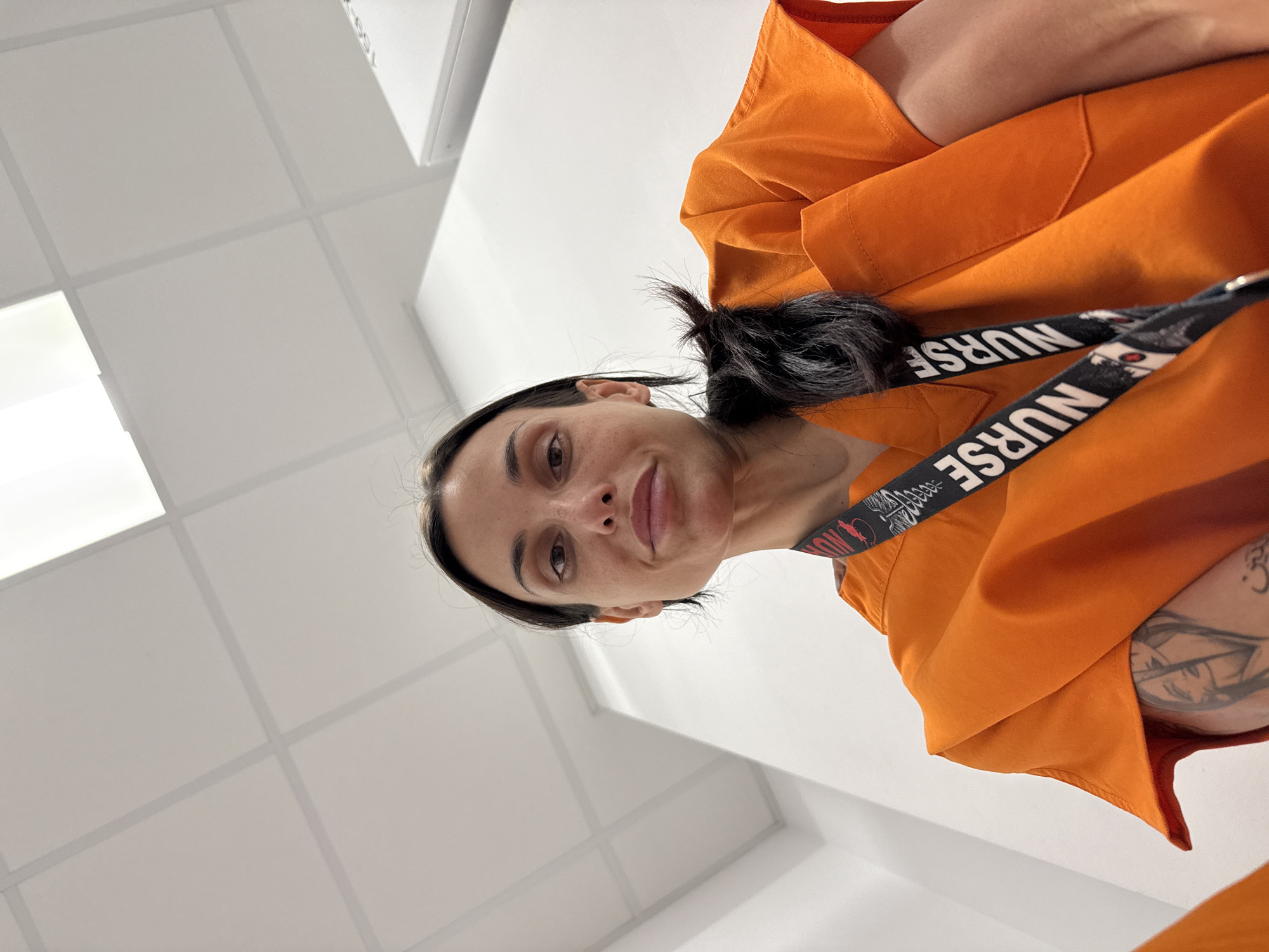 A woman with dark hair pulled into a ponytail and wearing an orange uniform with a "NURSE" lanyard around her neck is positioned in the lower center-right of the frame, looking directly at the camera. Her right arm, visible in the lower right corner, features a prominent tattoo. The background consists of a white tiled ceiling with recessed lighting. The location is unknown and there is no evidence to suggest it is in Pleven, Bulgaria.