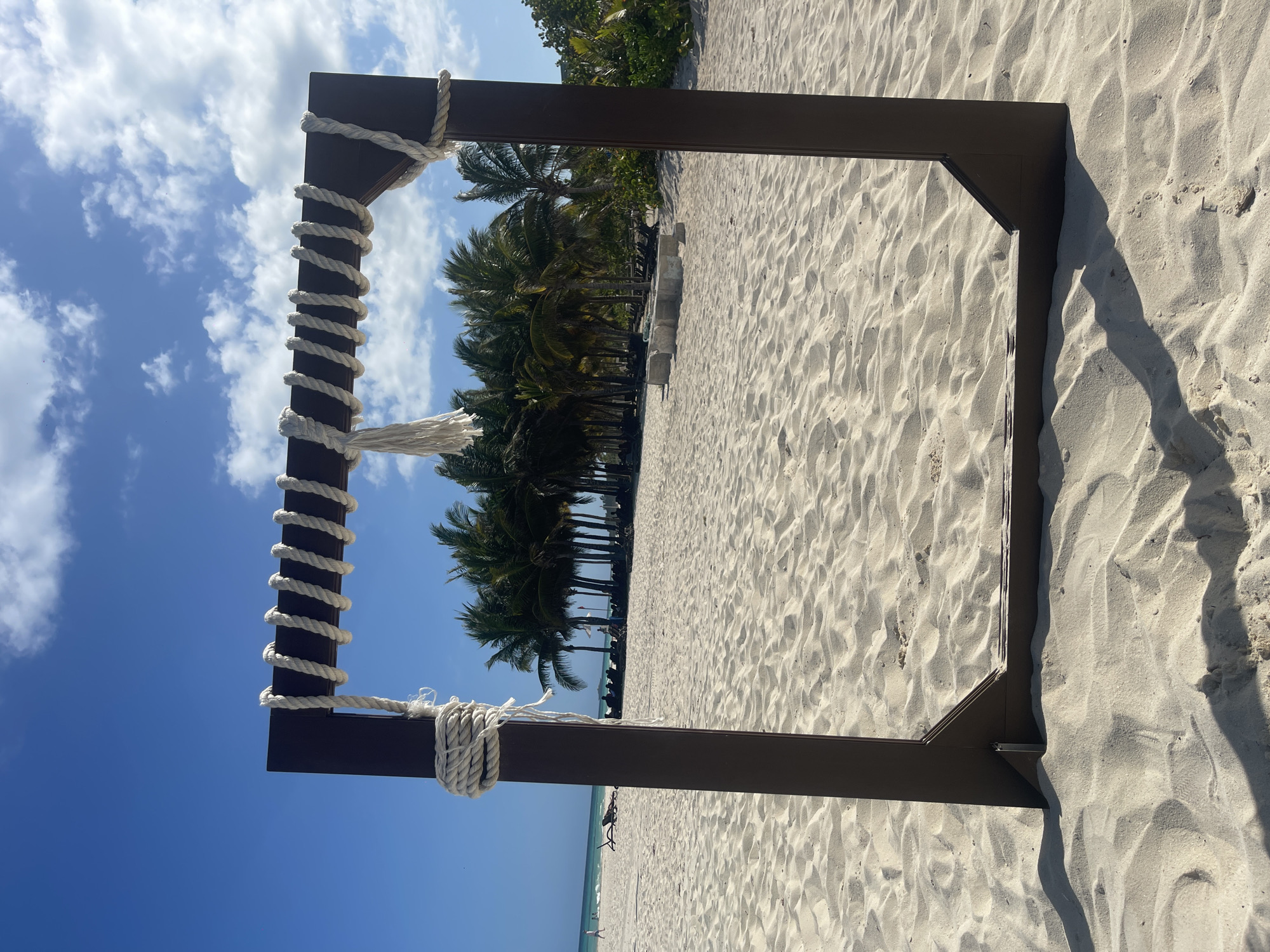The image shows a beach scene at Playa del Carmen, Mexico, viewed through a dark brown, wooden frame with a white rope wrapped around the left post. The frame is positioned on the beach, which is composed of light-colored sand. The background features palm trees against a blue sky with scattered clouds. The horizon line is visible with a glimpse of the ocean. A few people are present on the beach in the distance, but they are small and not easily discernible. The sunlight casts shadows on the sand, highlighting the texture.