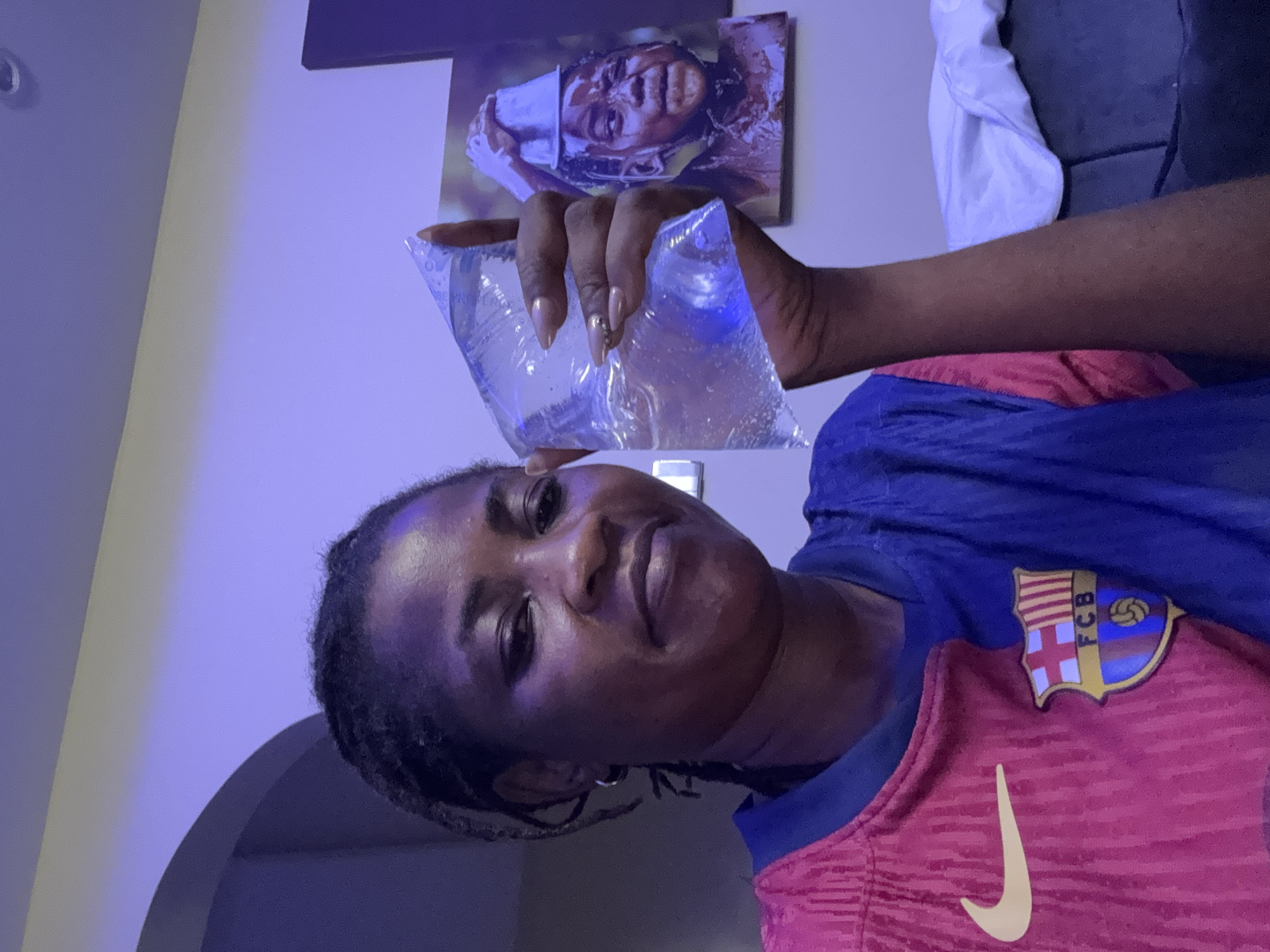 In a dimly lit room in Ajara, Nigeria, a woman with braided hair and wearing a red and blue soccer jersey is holding a clear plastic bag filled with water. Her nails are painted a light color and adorned with small rhinestones. In the background, a framed picture of a young person being splashed with water is visible. The ambient lighting casts a purple hue across the scene.