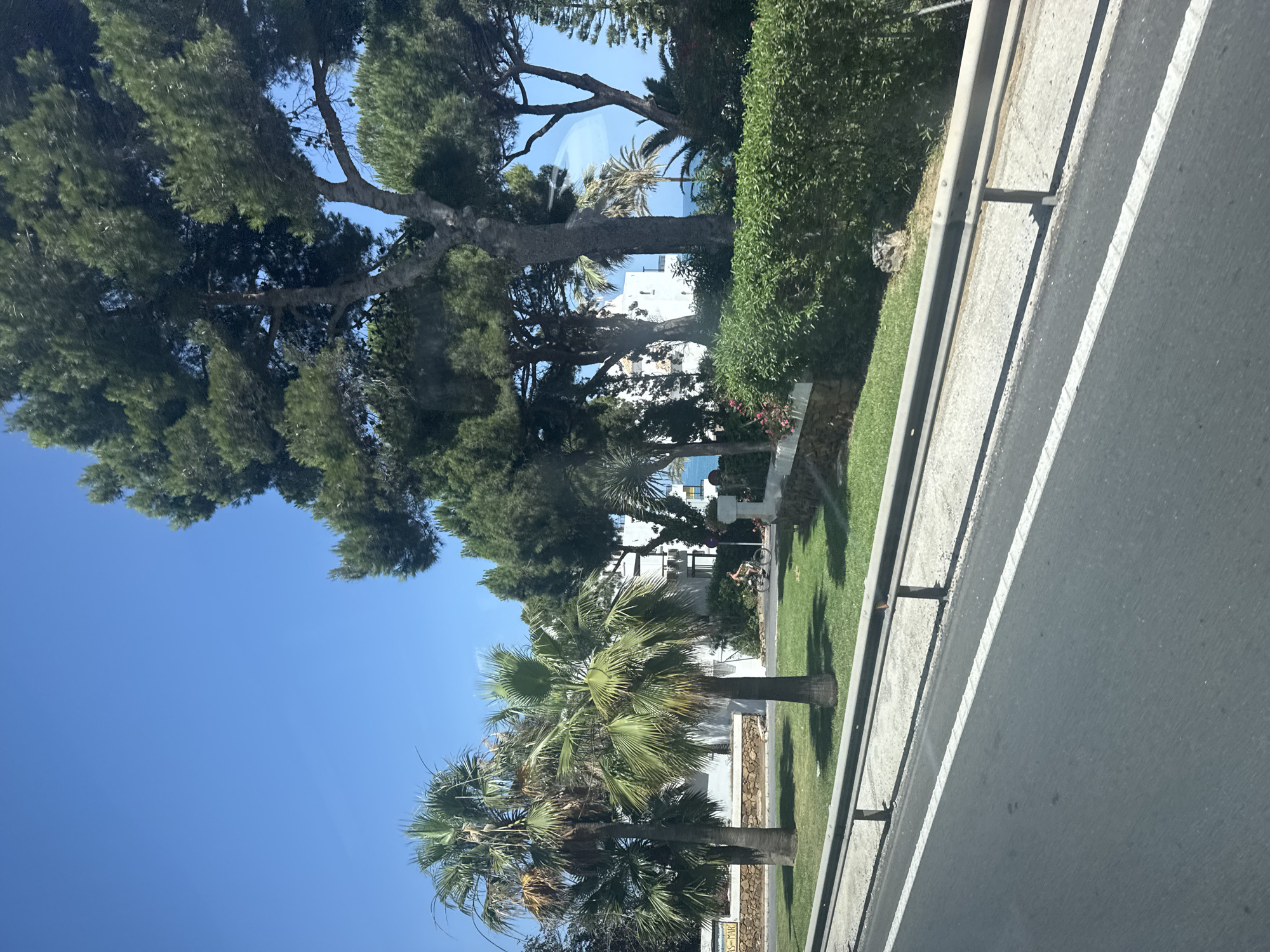 This image depicts a view from a vehicle traveling on a road in Mijas, Spain, on a clear, sunny day. The foreground shows the asphalt road with white lane markings and a metal guardrail. Beyond the guardrail is a strip of grass and a low stone wall. Behind this, a residential area is visible, characterized by white buildings with balconies, interspersed with lush green vegetation, including palm trees and tall pine trees. In the middle ground, a person is seen riding a bicycle on a path. Further in the distance, the blue sea is visible under a bright blue sky. The overall scene conveys a sense of a warm, coastal environment in a Spanish town.
