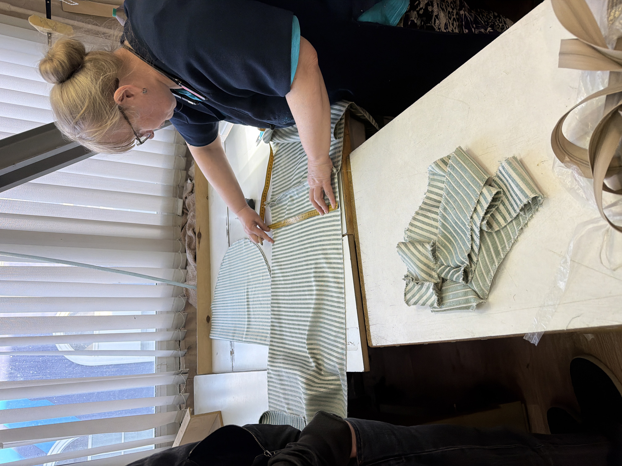 A person with light blonde hair pulled into a bun is standing over a white table, measuring a piece of striped fabric with a yellow measuring tape. The fabric appears to be partially cut into a curved shape. Other pieces of the same fabric are scattered on the table. A large zipper and a clear plastic bag are also on the table. The scene appears to be indoors, with blinds visible in the background. No evidence indicates that the location is in the city of Khimki, Russia.