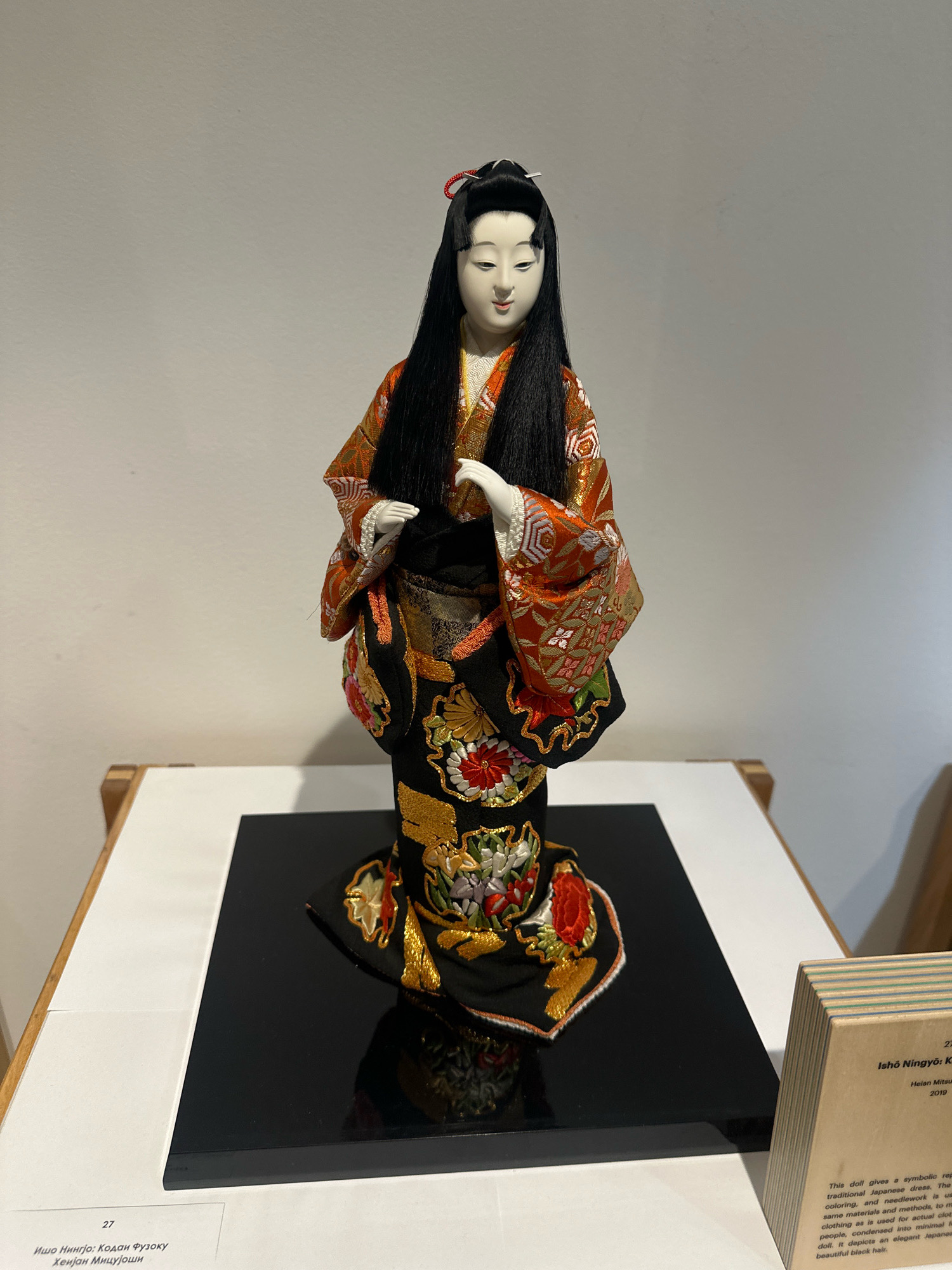 This is a full-shot, eye-level photograph of a traditional Japanese doll, likely an Ichimatsu doll, displayed on a glossy black pedestal. The doll is intricately dressed in a vibrant, multi-layered kimono. The kimono features a base of black fabric heavily embroidered with golden threads and floral motifs in red, pink, and white. The upper half of the kimono is a contrasting fiery orange with intricate patterns of white, gold, and possibly light green. The doll has a pale, porcelain-like face with delicately painted features, dark arched eyebrows, and red lips. Its most striking feature is its long, flowing black hair, which cascades down its back and shoulders. The doll's hands are posed gently in front of its body.
The doll is placed on a white surface, possibly a display table, against a plain, light-colored wall. To the right of the doll, a small, multi-layered wooden information plaque stands upright. The plaque has text printed on it, partially visible, which appears to be a description of the doll. The environment suggests an indoor exhibition or gallery space, possibly in Belgrade, Serbia, given the context of the request. The lighting is even and neutral, illuminating the subject clearly without harsh shadows.