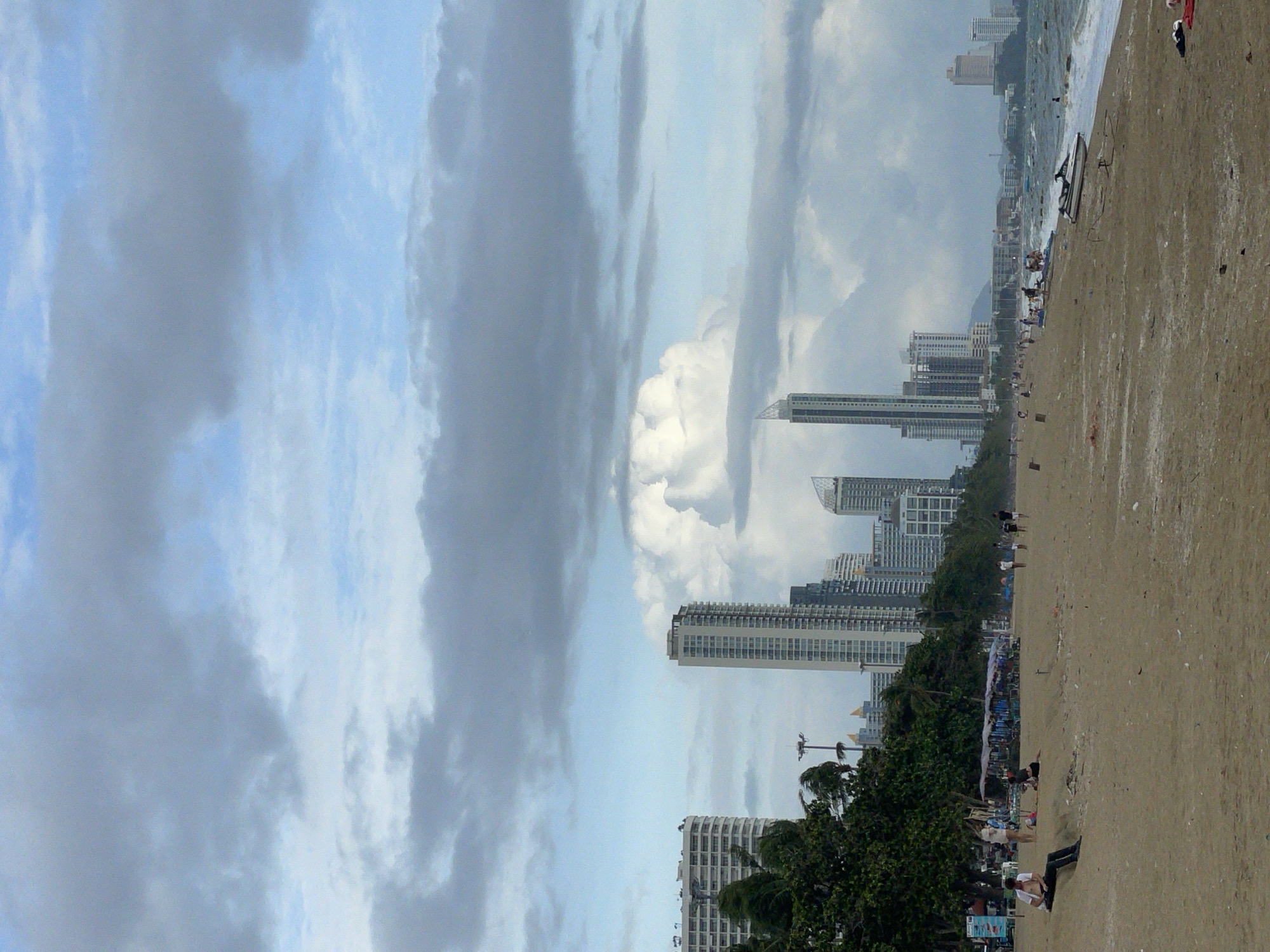 The image depicts a wide sandy beach in Pattaya City, Thailand, with the cityscape visible in the background. The sky is overcast with dramatic, dark clouds, suggesting a possibly stormy or moody atmosphere. Tall modern buildings, characteristic of a coastal city, dominate the skyline. The beach itself is sparsely populated, with a few individuals scattered about. In the foreground, a person is sitting on the sand, facing away from the camera, appearing relaxed. Further down the beach, a few more people are walking or gathered, and beach umbrellas are set up. The ocean is visible on the right, with waves lapping at the shore and a jet ski parked on the sand. The overall scene suggests a quiet moment on a busy beach, with the urban environment providing a striking backdrop to the natural elements.