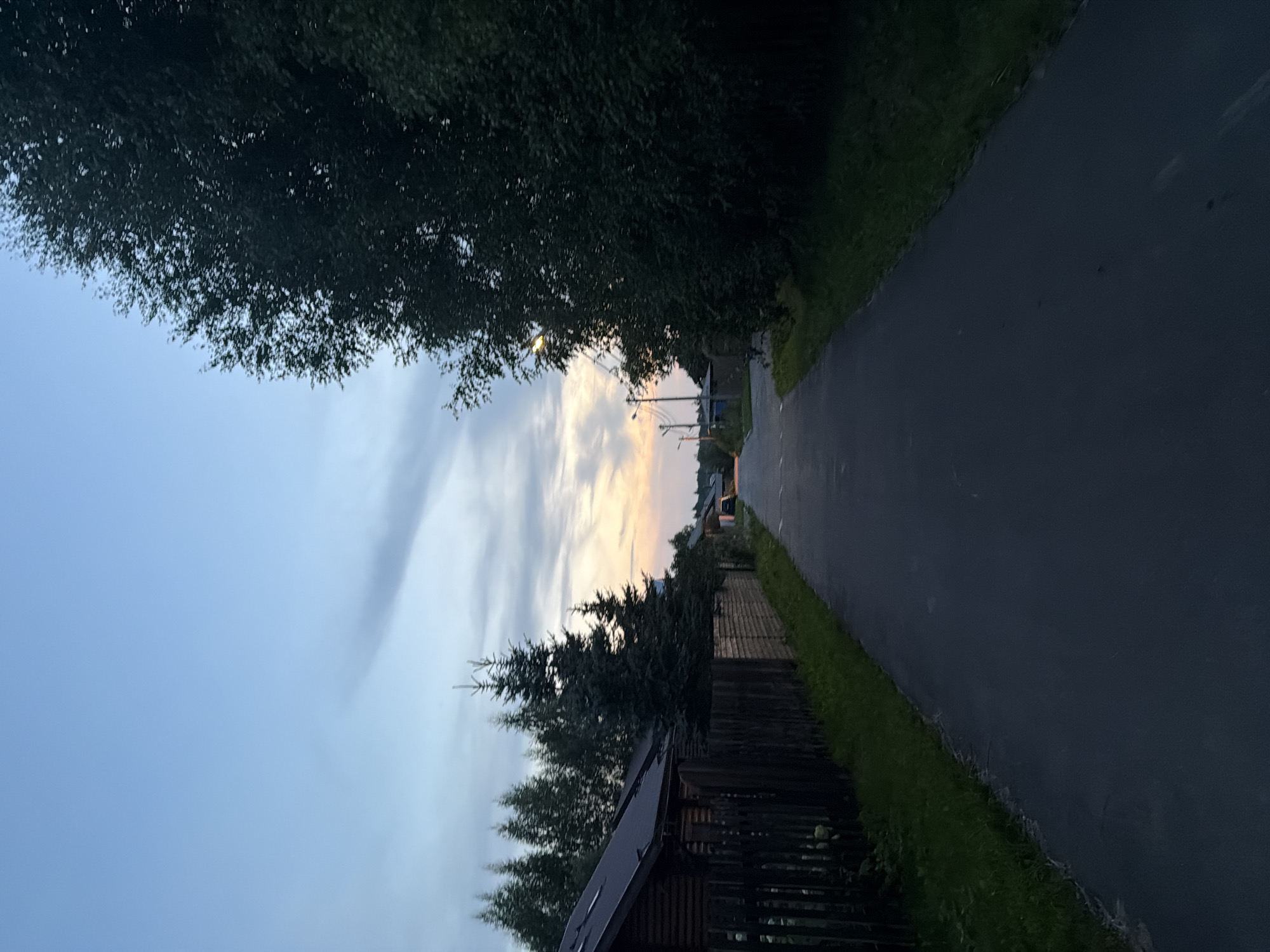 The image depicts a paved road in a suburban or rural setting, likely in Новорижские Ключи Кп, Russia. The road is flanked by dense trees on the right and a wooden fence and houses on the left. The sky is a gradient of blue and orange, suggesting either sunrise or sunset, with some clouds visible. Streetlights are present along the road. The overall impression is one of a quiet, tranquil evening. There are no people or animals visible in the scene. The time of day appears to be twilight.