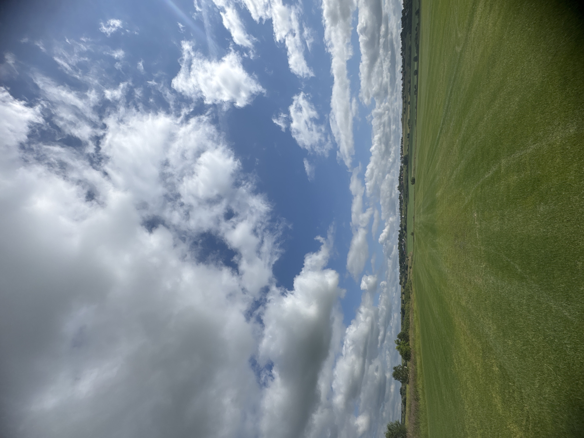 The image depicts a wide, well-maintained green field with rows of cut grass, suggesting it might be a park, golf course, or agricultural land. The field extends into the distance, meeting a line of trees and a low hill on the horizon. Scattered buildings are visible along the distant ridge, with a few houses and possibly some church spires. Above the landscape, a dynamic sky dominates the frame. Large, fluffy white clouds are interspersed with patches of bright blue sky, indicating a partly cloudy day with sunlight filtering through. The perspective is from ground level, looking slightly upwards towards the sky, emphasizing the vastness of both the field and the heavens. The overall impression is one of a serene, rural or semi-rural setting. The location is described as being in the city of Kempelevo, Russia.