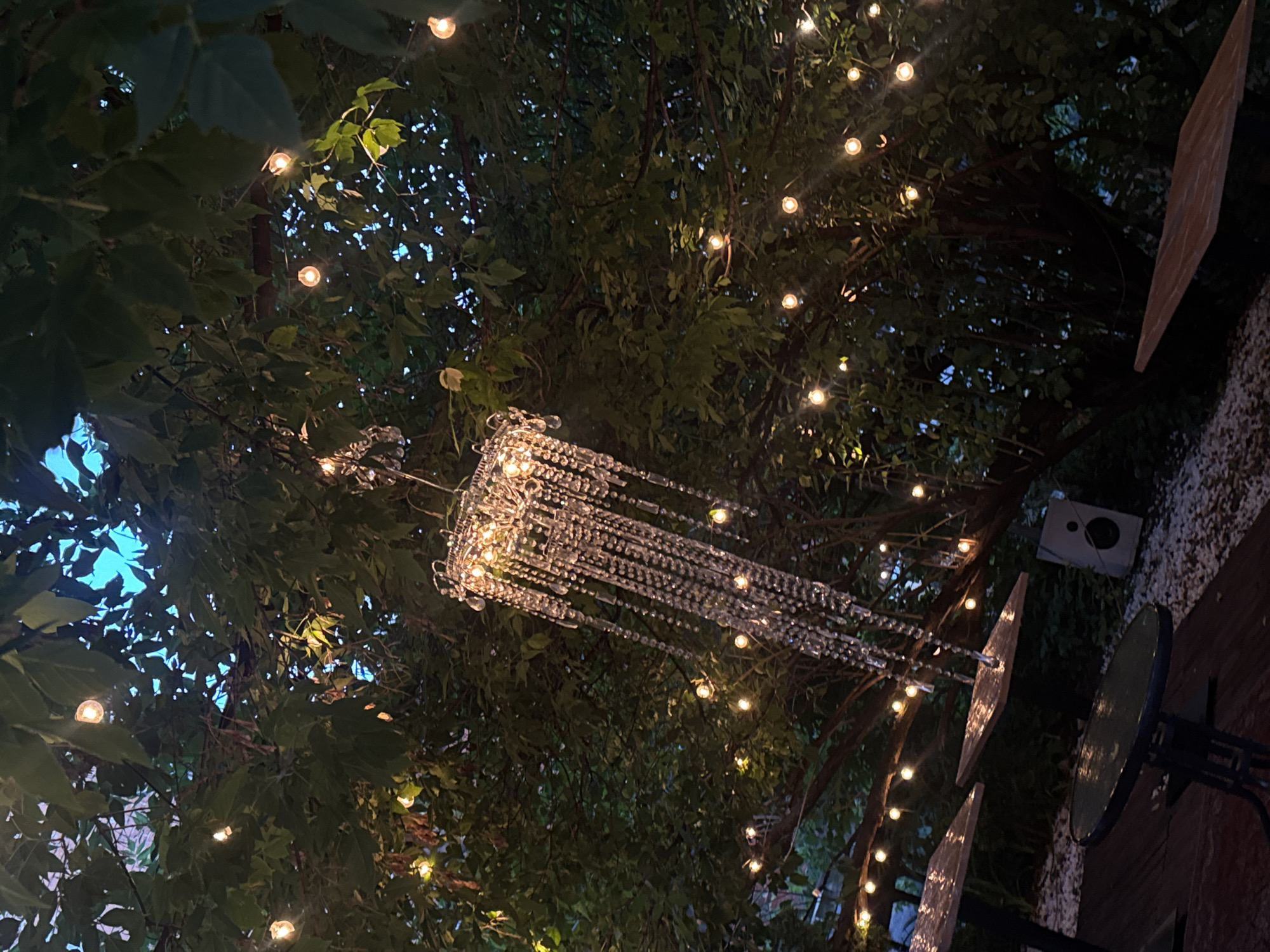 The image depicts an outdoor seating area, likely a cafe or restaurant patio, adorned with string lights and a crystal chandelier. The scene is set at dusk or nighttime, with the soft glow of the lights illuminating the lush green foliage overhead. Several small tables with dark bases and reflective tabletops are visible, some with a reddish-brown hue, suggesting a wooden or stained surface. In the lower left, a round table with a dark base and a glass top is in focus. To the right of the tables, a white speaker box rests on a bed of white pebbles. The chandelier, hanging from the branches of a large tree, is elaborate with numerous cascading crystal elements that catch and refract the light. The overall ambiance is festive and inviting, hinting at a charming urban setting in Nizhny Novgorod, Russia.