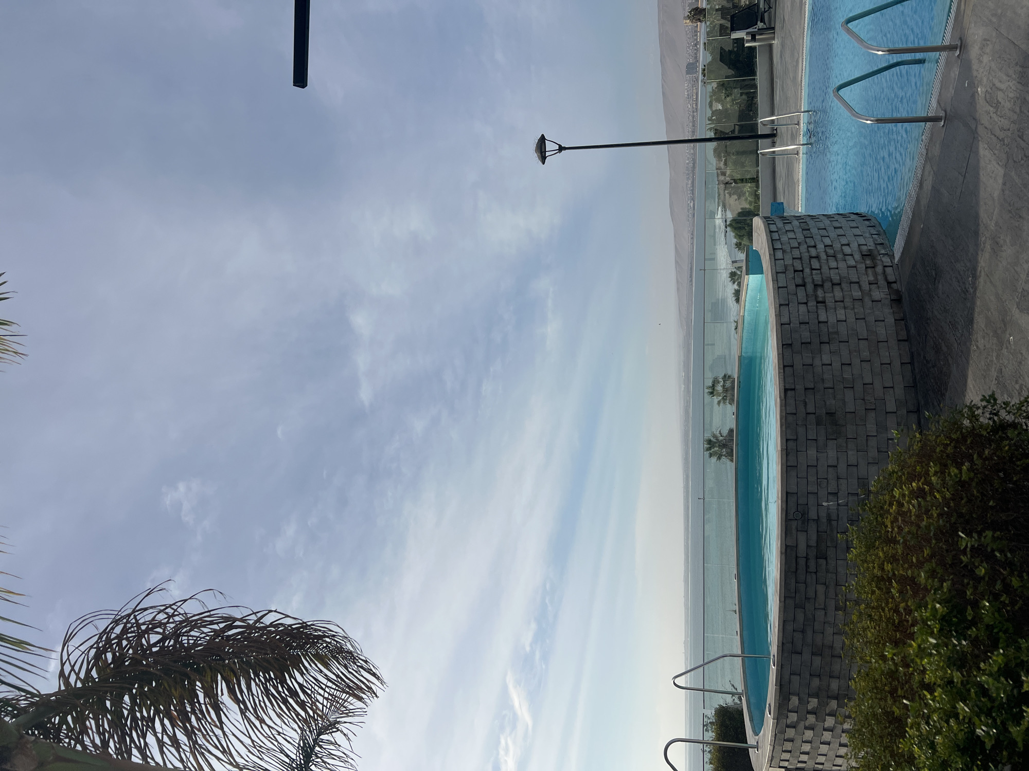 The image shows an outdoor swimming pool area in Arica, Chile. Two swimming pools, one rectangular and one curved with a brick border, overlook the ocean. A glass barrier separates the pool area from the view. A palm tree branch is visible in the lower left corner. A lamppost stands between the pools. The sky is partly cloudy.