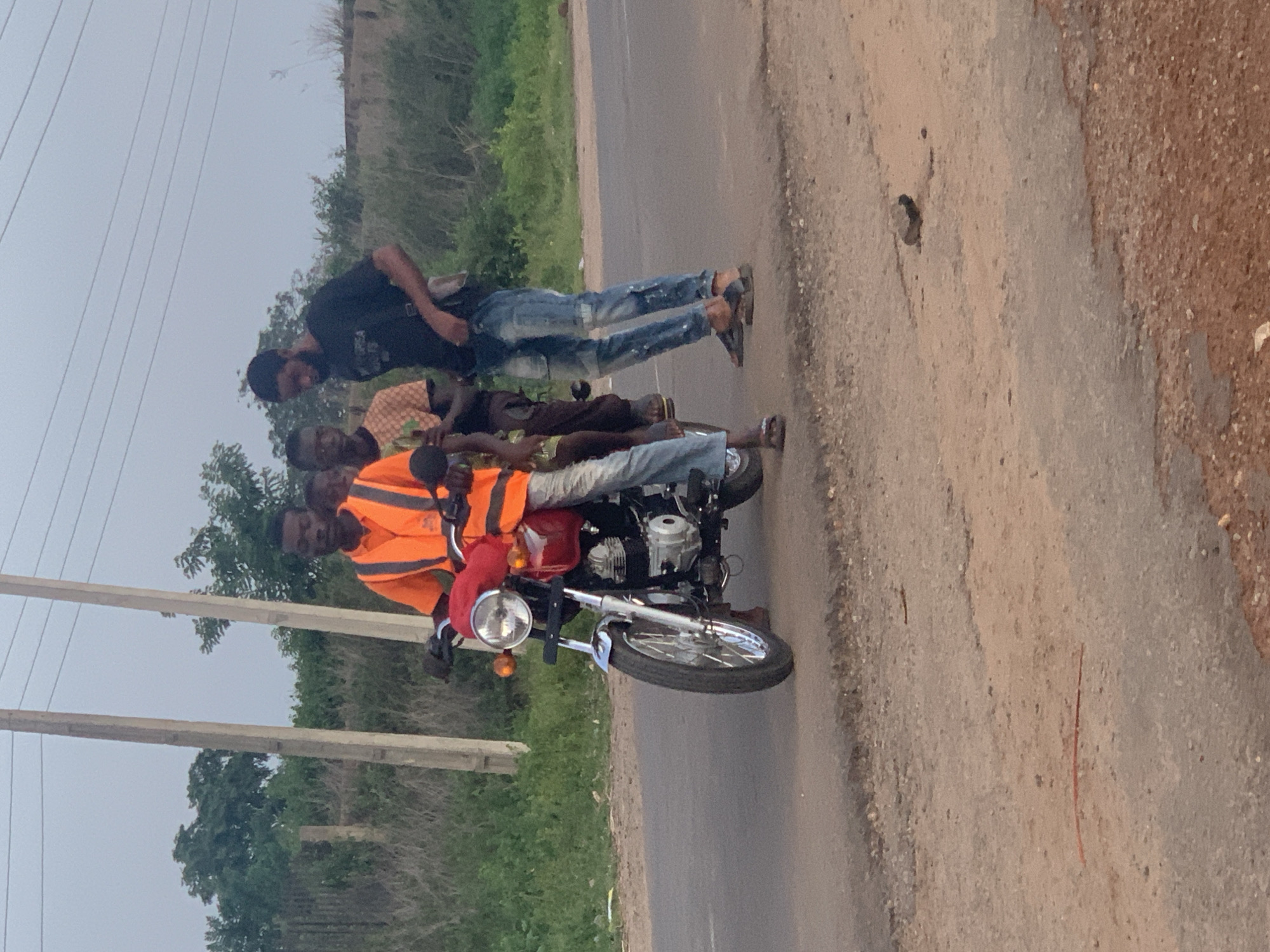 In Gbeleko, Nigeria, four people are on a red and chrome motorcycle on a paved road. The rider wears a reflective orange safety vest. Two passengers are seated behind the driver, and a fourth person walks alongside the motorcycle to the right. The surrounding environment includes lush greenery and utility poles.