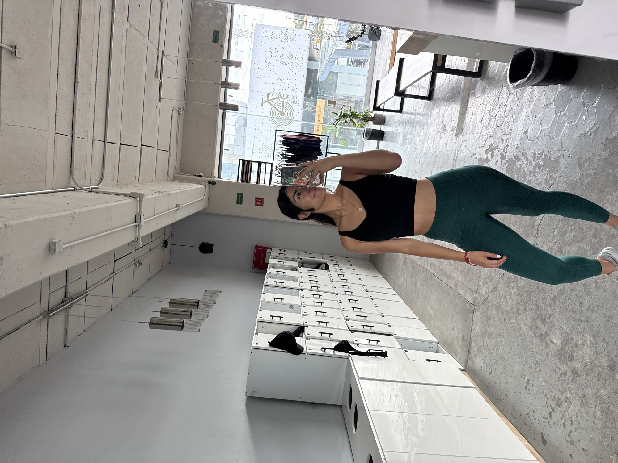 The image shows a woman taking a selfie in a fitness studio located in Puebla, Mexico. She is wearing a black top and green leggings. Behind her is a row of white lockers with black hats on top. The floor is concrete. A large window provides natural light and offers a view of buildings outside. The gym's logo is visible on the window.