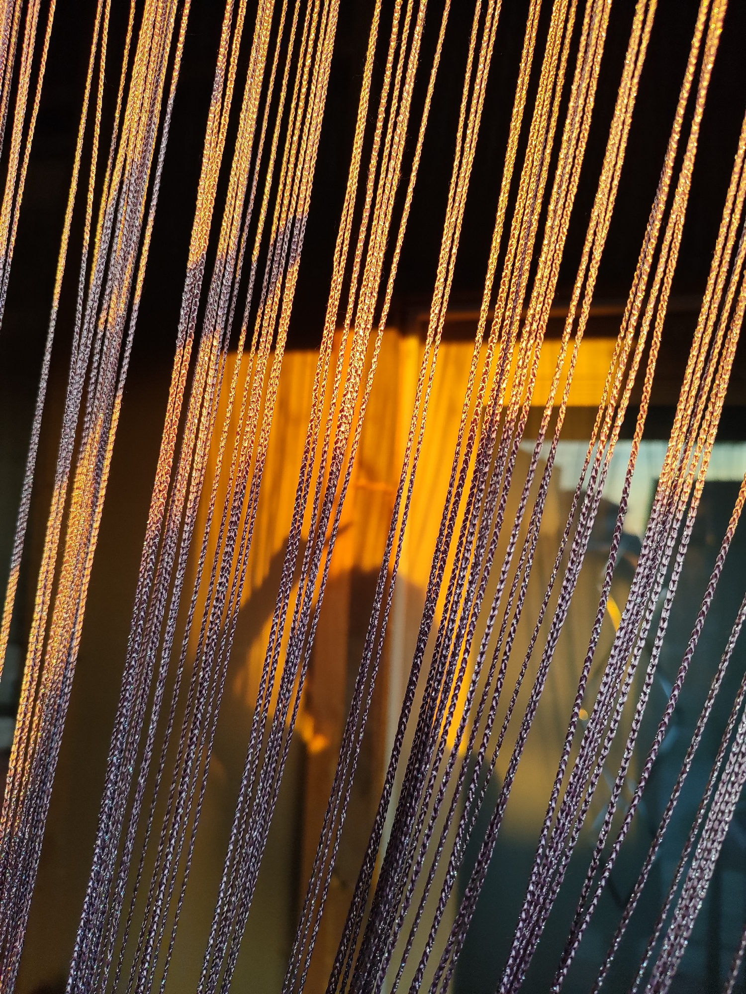 This image captures a close-up view of a string curtain illuminated by warm, golden sunlight. The curtain is composed of numerous thin, braided strands, varying in color from dark purple to a lighter, almost silvery hue, with some strands reflecting the sunlight with a metallic sheen. The sunlight creates distinct shadows on the wall behind the curtain, suggesting it is positioned in front of a window or doorway. The warm light indicates it is likely either sunrise or sunset. The background wall has a rough, textured appearance and is painted a muted yellow or ochre color. The overall mood is serene and peaceful, with the play of light and shadow adding depth and visual interest to the simple subject matter. No people, text, or specific location cues beyond the general hint of a domestic setting are visible. The image is likely taken in Marusino, Russia, as provided.