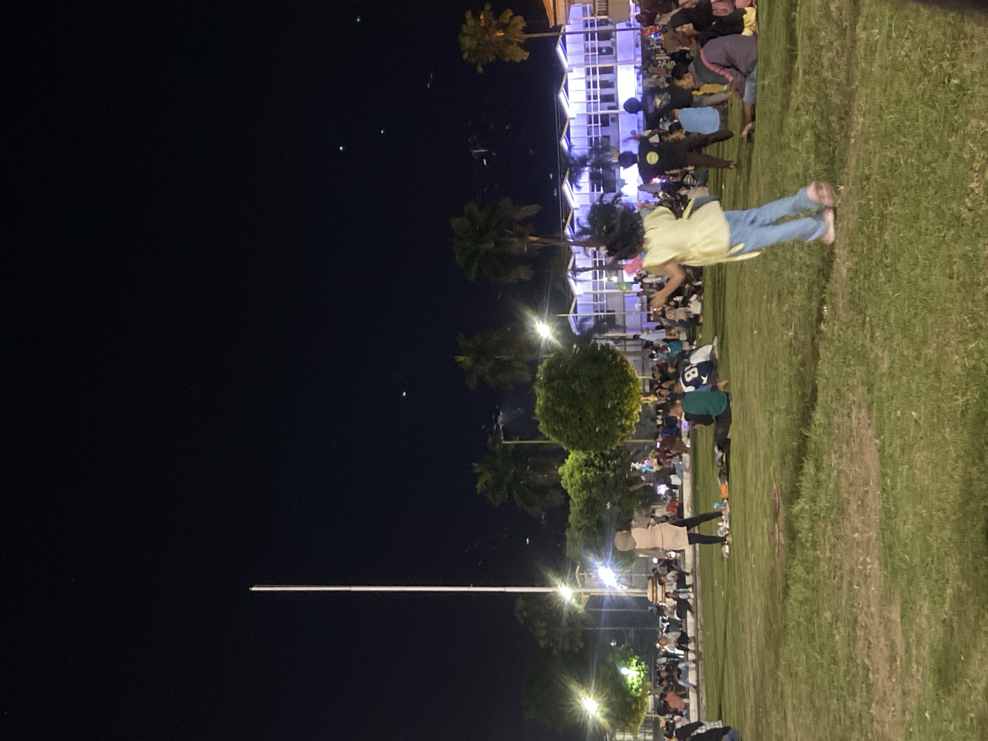 The image depicts a nighttime scene in what appears to be a park or public space in Jember, Indonesia. There is a large crowd gathered, with people of various ages and attire visible. Some individuals are standing on a grassy area to the right of the frame. A few people are actively engaged in running. A line of street lights illuminates the area. Palm trees and other greenery are interspersed among the buildings and crowds. The sky is dark, suggesting it is late in the evening.