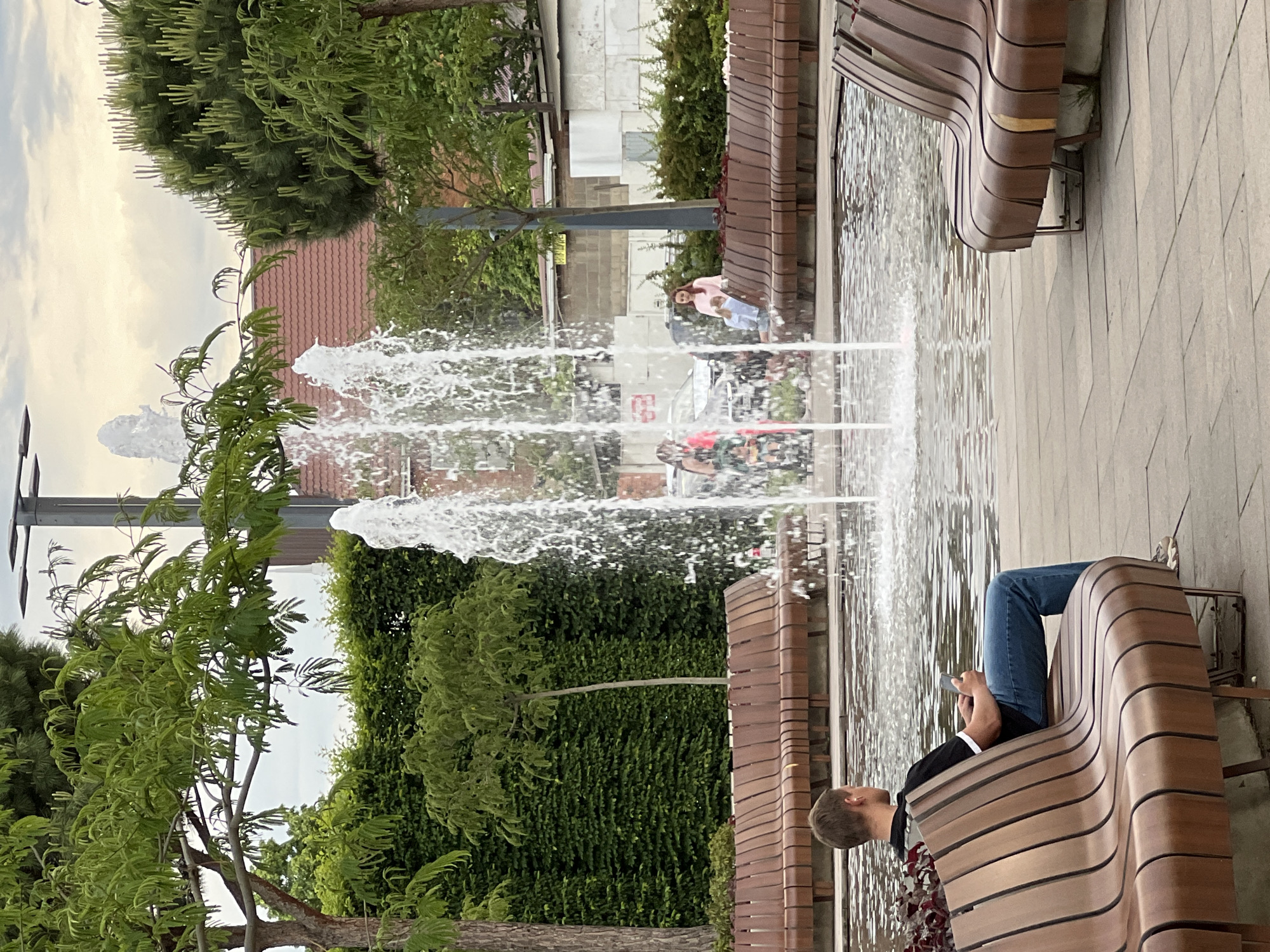 The image depicts a serene urban park or public square in Krasnodar, Russia, featuring a modern linear fountain as its central element. The scene is captured during daytime under an overcast sky, providing soft, diffused lighting.
In the foreground, a person, likely a man, is relaxing on a contemporary, curved wooden slatted bench. He is wearing a black shirt and blue jeans, with his legs propped up on the bench while he engages with his mobile phone. The bench itself is part of a series of similar benches bordering the fountain's water channel.
The fountain consists of several upward-shooting water jets, creating dynamic splashes and reflections on the water's surface. In the mid-ground, near the fountain, a woman in a pink top and light blue pants stands observing the area. Other indistinct figures can be seen further in the background, interacting with the space around the fountain.
The environment is lush with greenery, including several trees with vibrant leaves and a dense wall of dark green foliage or ivy. Beyond the trees and fountain, buildings with reddish-brown tiled roofs and lighter-colored walls are visible in the background, suggesting a city setting.
Visible text is scarce and mostly illegible due to blurriness and distance. There appears to be a small, blurry yellow sign in the mid-ground that could be a "TAXI" sign, and some indistinct red letters/numbers, possibly "59" or "5G," on a surface behind the woman. The overall atmosphere is one of leisure and calm in a well-maintained public space.