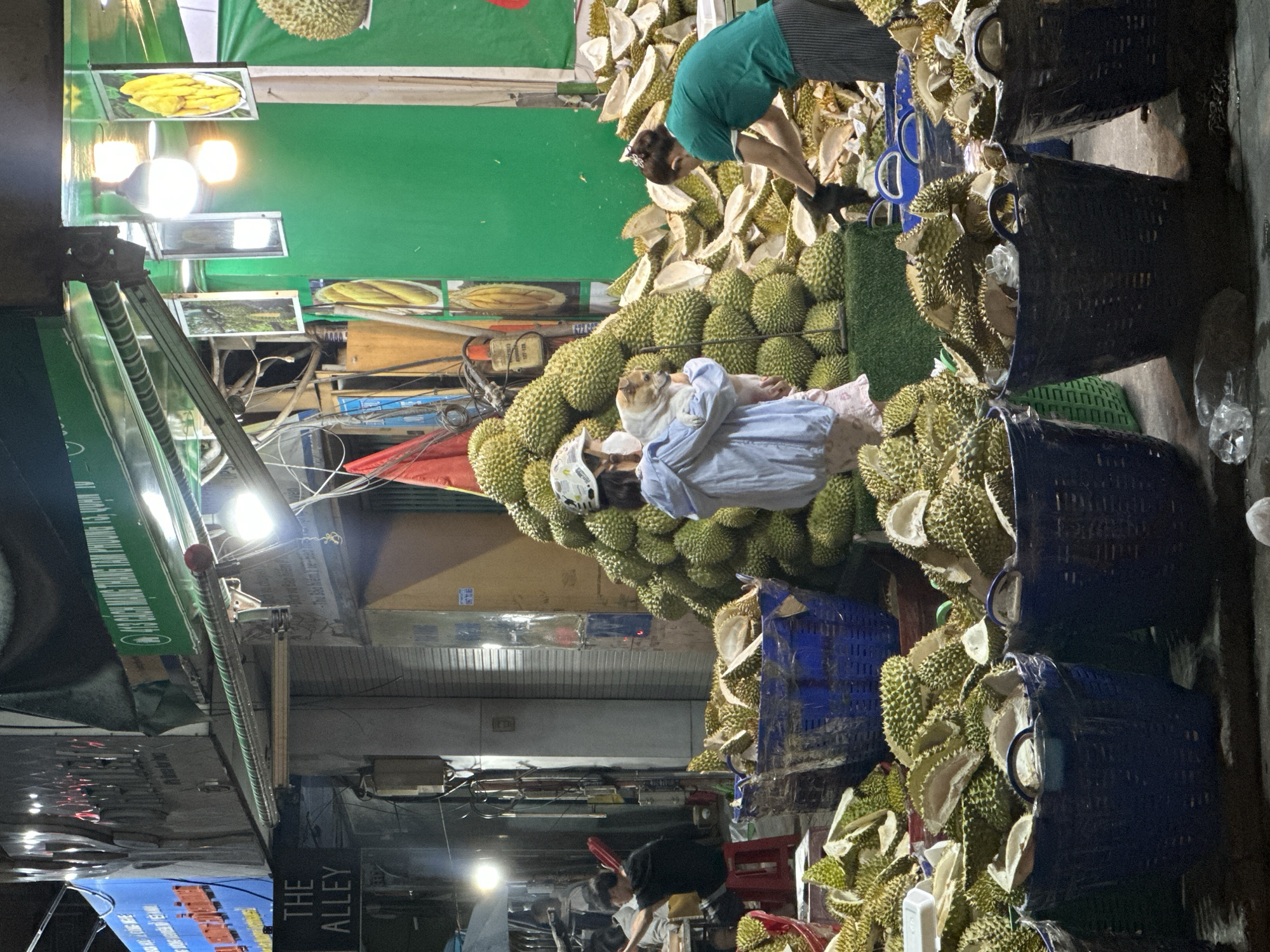 The image shows a street scene in Thủ Đức, Vietnam, dominated by a durian fruit stall. A large pile of durians is the central focus, with a woman in a green shirt and striped pants reaching over the pile. A person in a light blue shirt is resting on top of the durian pile, partially covered by a blanket. Baskets filled with durians and durian shells are stacked nearby. Street signs are visible in the background and to the left of the frame, and there are a few other people further down the street near the bottom of the image. A store sign reads, "THE ALLEY". Overhead lights illuminate the durian stall and the surrounding area.