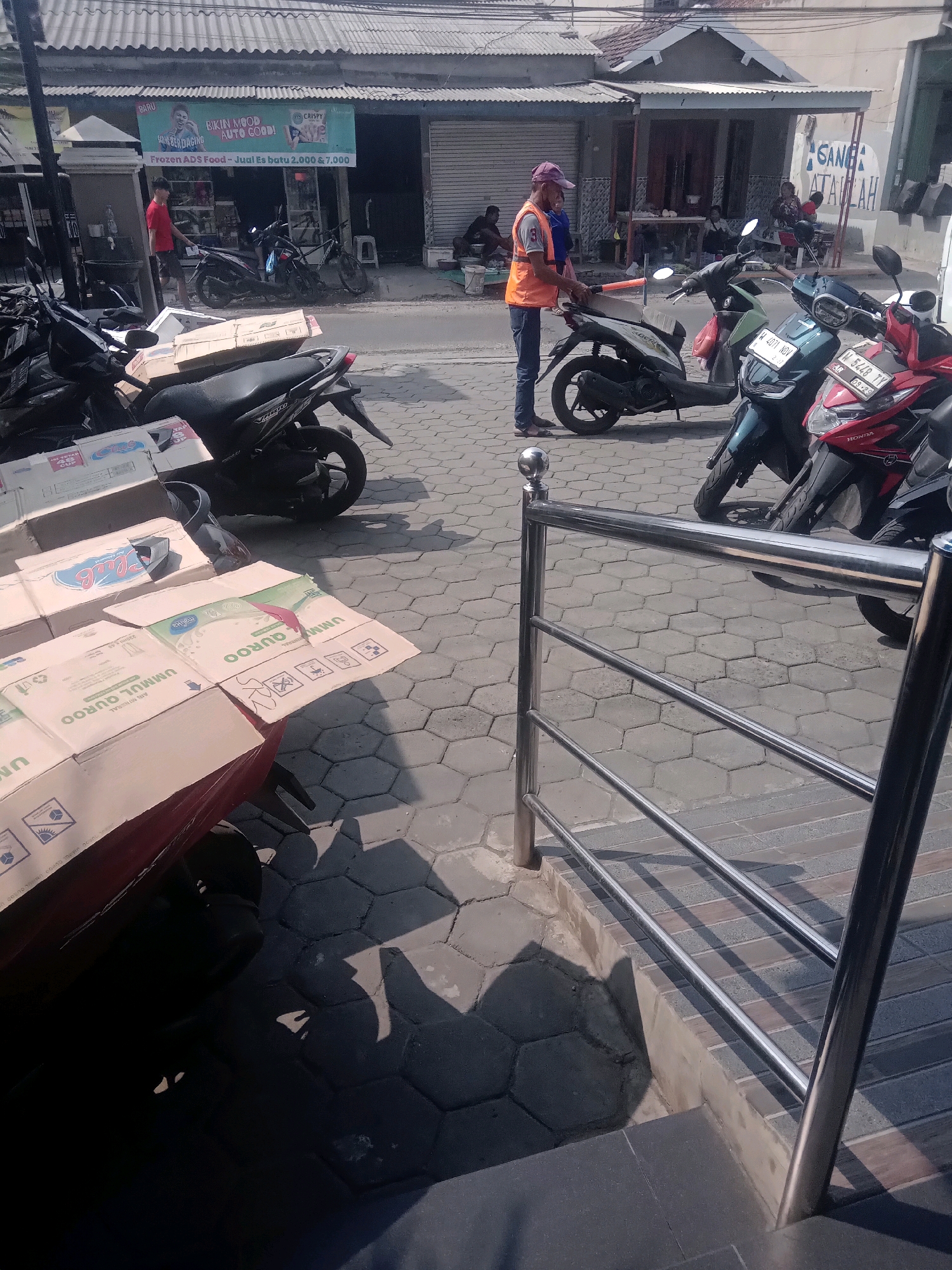 The image shows a street scene in front of a commercial establishment in Pepelegi, Indonesia. In the foreground, several motorcycles are parked on a cobblestone pavement. Some cardboard boxes, possibly containing goods, are stacked on the back of a red motorcycle. A metal railing runs along the edge of a raised area. In the mid-ground, a man wearing an orange vest and a purple cap is interacting with a motorcycle. Further back, more motorcycles are parked, and a few people are visible in front of a storefront with a corrugated metal roof. The storefront displays promotional signs. The overall environment suggests a commercial street with an active street life.