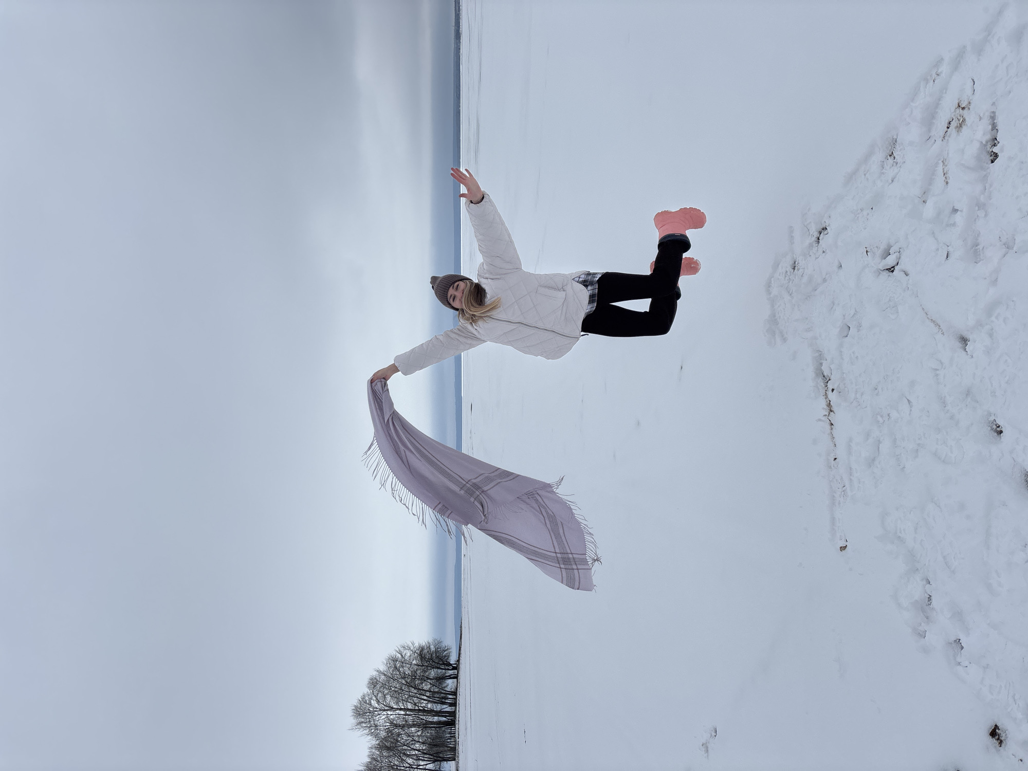 A person stands in a snow-covered landscape, holding a gray, fringed scarf in their left hand. They wear a white quilted jacket, black pants, a brown beanie, and pink boots. Their arms are outstretched, as if in motion. The background features a body of water or sky with a horizon line, and leafless trees are visible at the bottom. The setting could be the city of Chaikovsky, Russia. Patches of disturbed snow can be seen in the bottom right corner. The image is rotated 90 degrees clockwise.