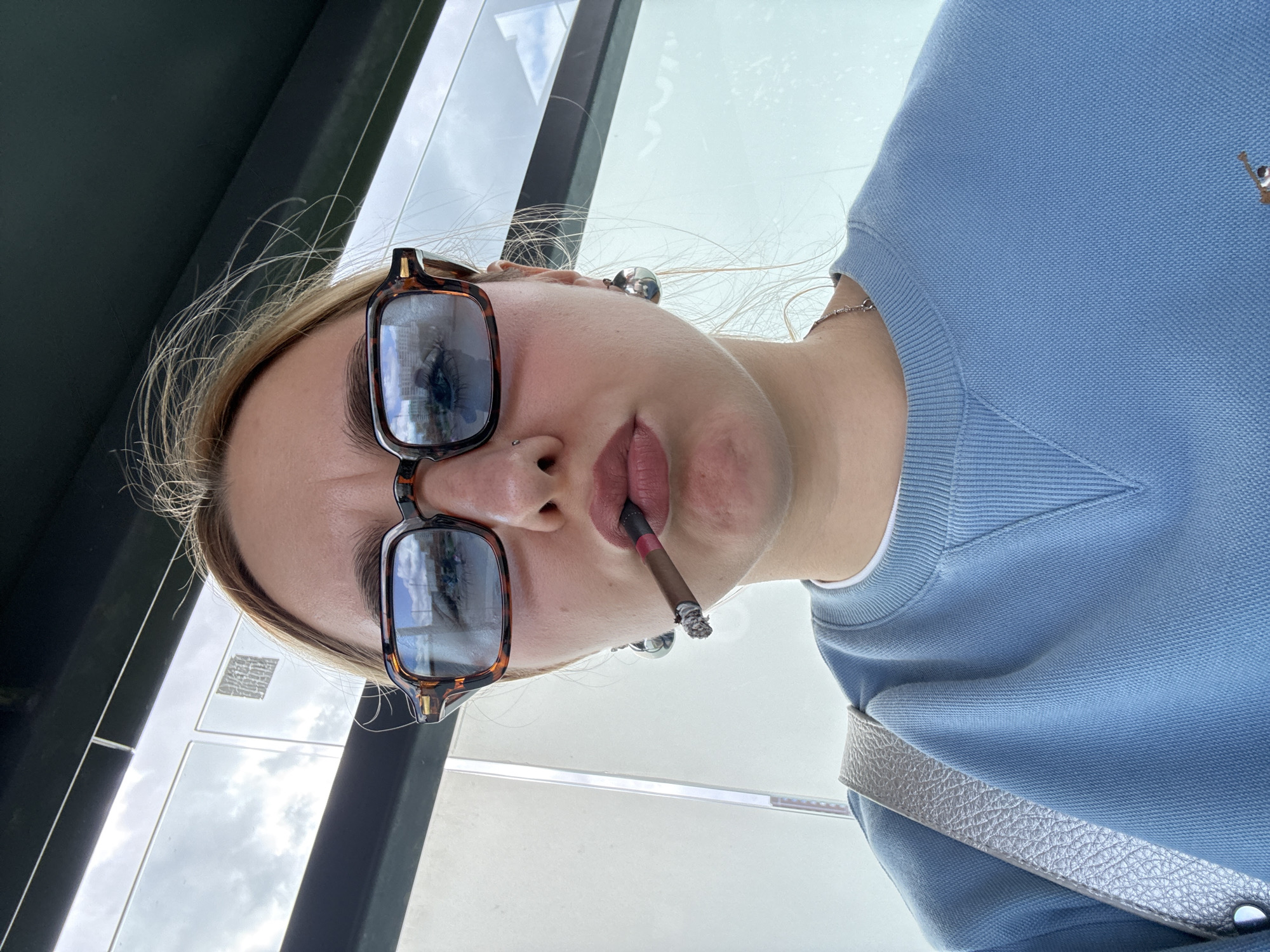A young woman with blonde hair and a nose piercing is holding a lit cigarette in her mouth, wearing tortoiseshell glasses and a blue sweater with a small logo, possibly standing at a bus stop. The reflection in her glasses shows urban architecture, suggesting a city environment. No specific landmarks identify it as Minsk, Belarus, but the general architecture is consistent with urban areas.
