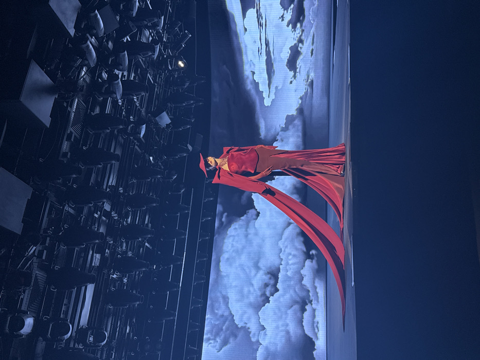 A singer stands on a stage in a long red dress with flowing fabric. Behind her is a screen displaying a cloudy sky background. The scene is lit with blue and white lights. To the left, a large grid of stage lighting equipment is visible. The overall impression is a theatrical performance.