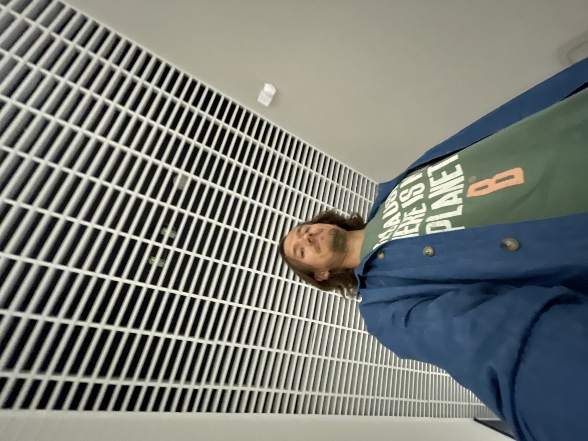 A man with long brown hair stands in front of a white, gridded wall. He is wearing a green t-shirt with white lettering and a blue jacket. The location is Unknown, Unknown.