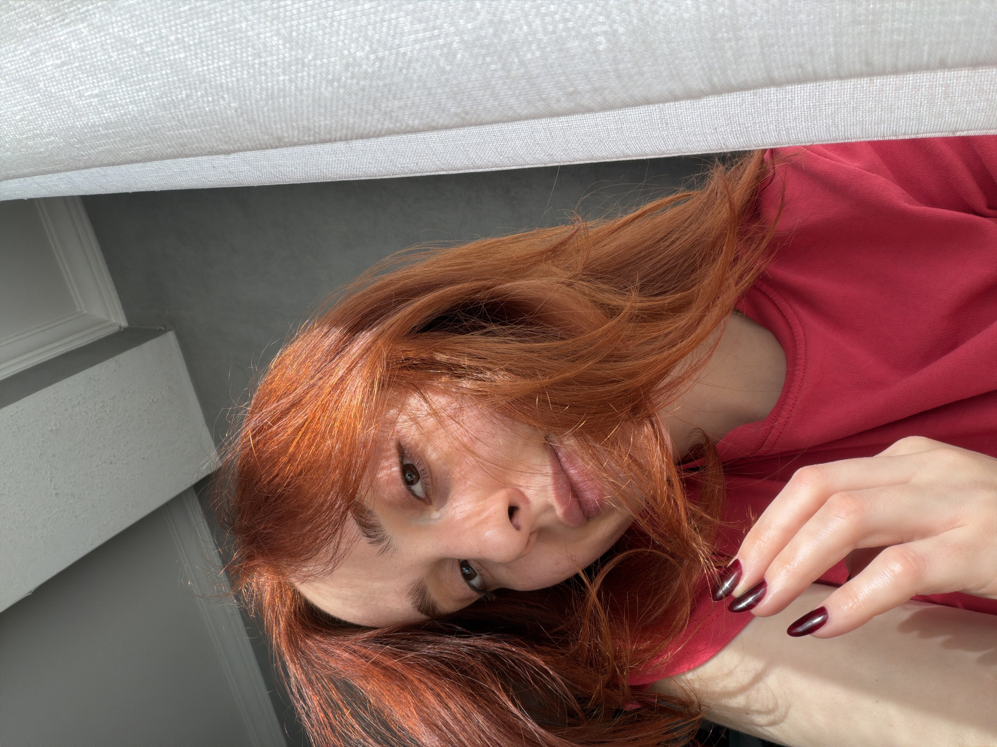 A woman with auburn hair is lying down, looking up towards the camera. She is wearing a red shirt. Part of a white upholstered object is visible above her head. Her right hand is raised slightly, with dark red nail polish. The background includes a gray wall and white trim. No specific location in Tbilisi, Georgia, can be determined from the image.