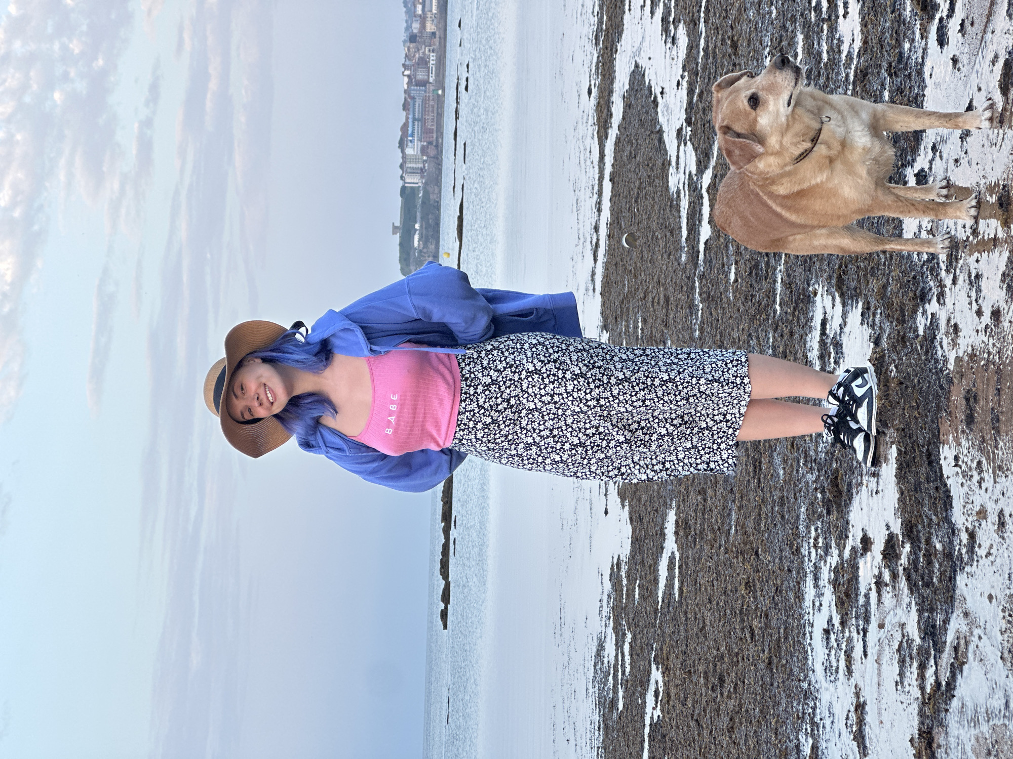 On a flat, algae-covered shore in Gijón, Spain, a woman in a brown straw hat and blue-tinted hair smiles while standing next to a golden Labrador dog. She wears a pink tank top with the word "BABE" on it and a blue hoodie, a black and white floral skirt, and black and white sneakers. The dog looks upwards. Behind them, the sea stretches towards the city skyline in the background. The sky is overcast.