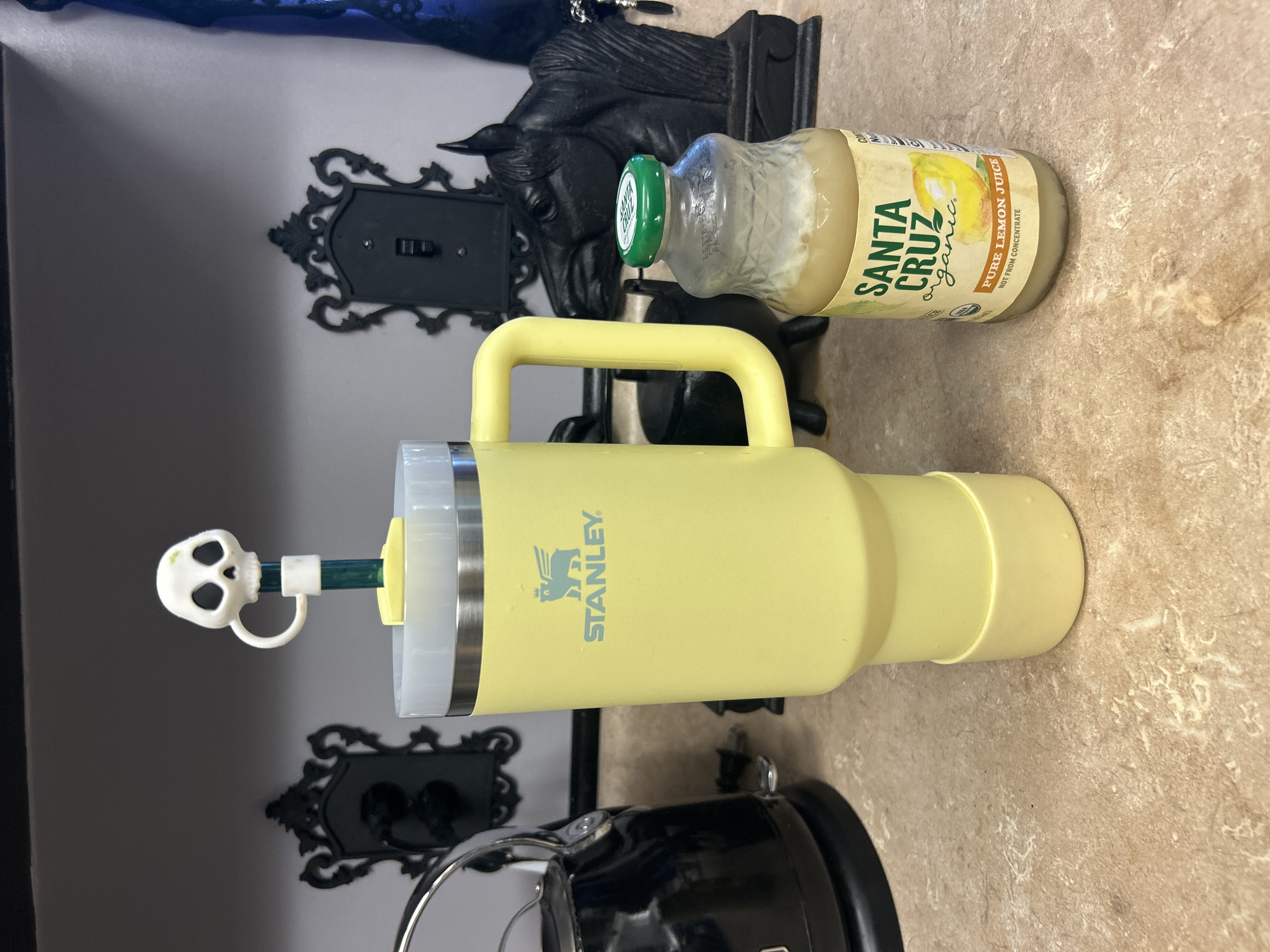 This indoor shot shows a yellow Stanley tumbler with a skull-shaped straw topper, a bottle of Santa Cruz Organic Pure Lemon Juice, and decorative black metal objects on a countertop. The countertop has a light brown, speckled pattern. Behind the tumbler and juice are two ornate black metal wall decorations, one of which appears to be a light switch plate. In the lower part of the frame, a portion of a black kitchen appliance with chrome trim is visible. The location is Cape Coral, United States of America, inferred as the Santa Cruz organic product is more common to the USA, and the general interior style and commonness of Cape Coral, United States of America.