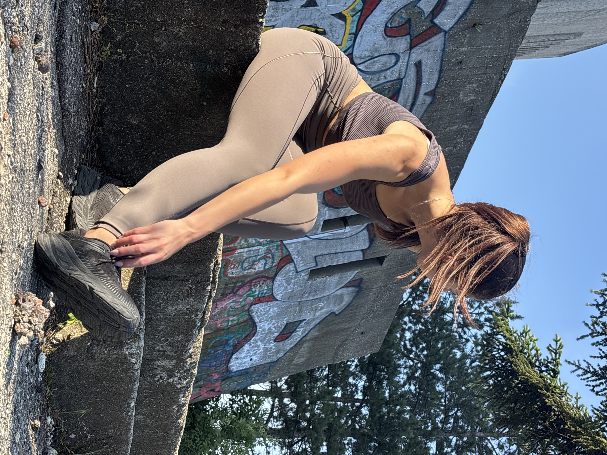 A medium shot, rotated ninety degrees clockwise, captures a person engaged in a stretching or reaching pose outdoors. The main subject is a woman with brown hair, dressed in grey/taupe athletic leggings and a dark grey ribbed sports bra or crop top. She wears dark athletic shoes, possibly Hoka, and a delicate silver-colored necklace is visible around her neck. Her body is bent forward from the waist, with one hand reaching towards her foot, suggesting she is adjusting her shoe or performing a stretch.
The setting appears to be an urban or semi-urban outdoor space, possibly in Varna, Bulgaria, as indicated by the context. The person is positioned against a textured, rough concrete structure, which could be steps, a wall, or a barrier. This concrete surface is adorned with colorful graffiti, featuring abstract shapes and lines in white, red, blue, and green, suggesting a public or neglected area.
In the background, beyond the concrete structure, a clear blue sky is visible, indicating fair weather. Lush green coniferous trees are also prominent, adding a natural element to the scene. The bright, direct sunlight suggests it is daytime, casting noticeable shadows. No legible text is visible in the graffiti or elsewhere in the image.