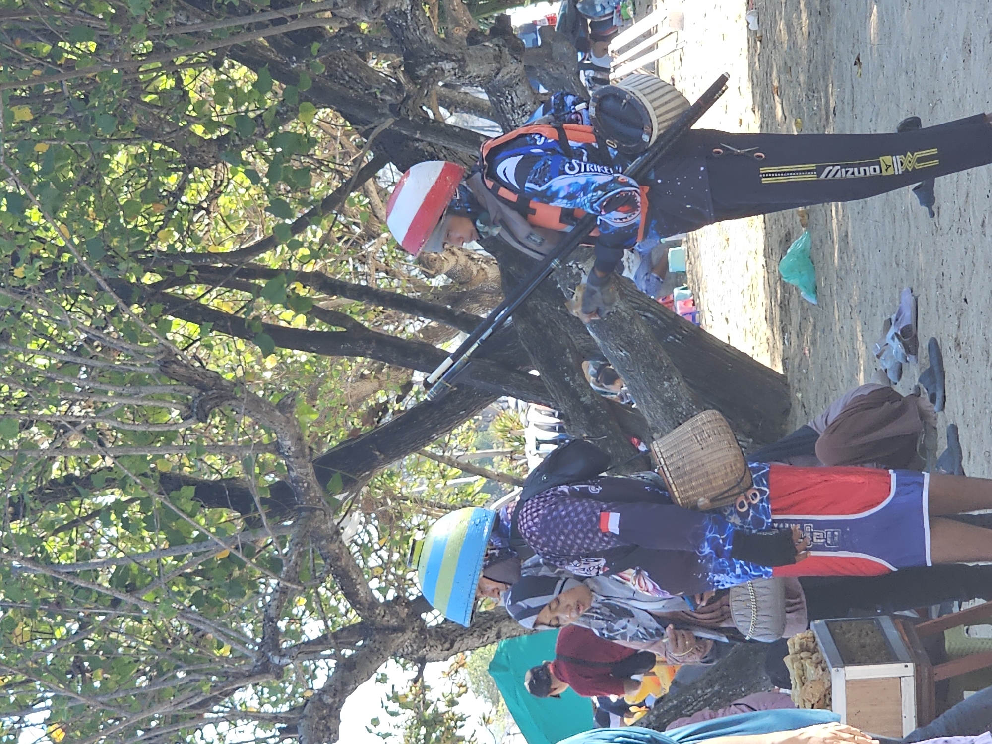 The image shows a group of people on a sandy beach, possibly near Denpasar, Indonesia. The scene appears to be outdoors during the daytime, with bright sunlight casting shadows and illuminating the sand and trees.
The primary focus is on two individuals in the foreground, dressed in athletic or specialized attire, suggesting an activity like fishing or spearfishing. The person in the upper portion of the frame wears a blue and black suit with a life vest and a red and white striped helmet. They are holding what appears to be a speargun. The person in the lower portion of the frame wears a similar outfit, including a colorful helmet with blue, yellow, and green stripes, and a patterned long-sleeved shirt. They are also positioned as if engaged in an activity, with a basket attached to their back.
The background is characterized by the sandy beach, with some vegetation and trees providing shade. There are also other people visible in the background, some appearing to be resting or observing. Scattered objects like bags and what look like fishing gear or accessories are present on the sand.
Notable details include the clothing and equipment, which point towards a water-based activity. The bright sunlight and clear visibility suggest fair weather. The overall atmosphere seems relaxed and casual, typical of a beach setting. Visible text on the legwear of the person in the upper frame clearly reads "MIZUNO".