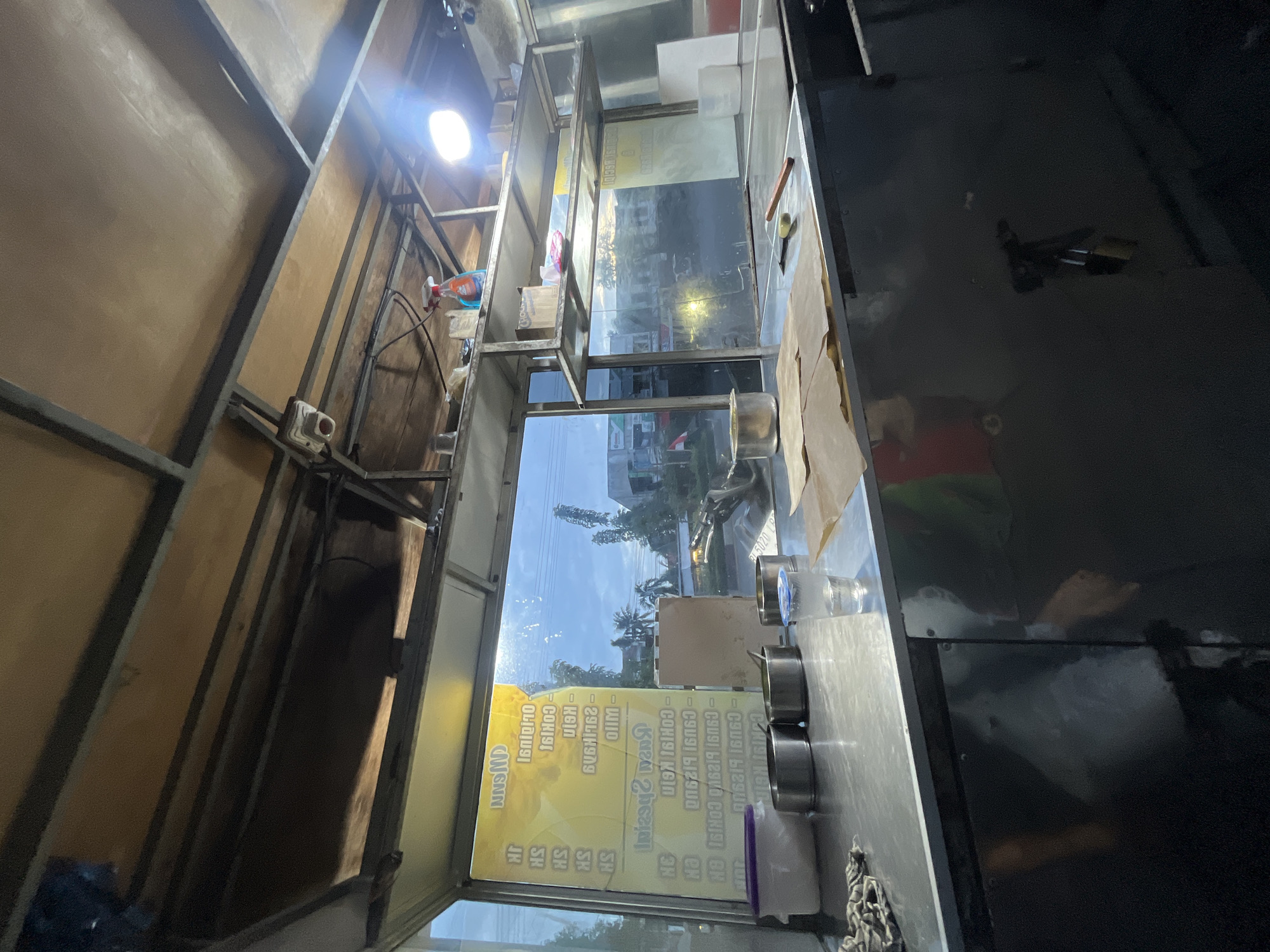 The image shows the interior of a food stall in Banda Aceh, Indonesia. The stall appears to be open for business, with the serving window facing outwards. Inside the stall, the counter is made of stainless steel and is cluttered with various items, including metal containers, a glass of water, a rag, and some brown paper. There is a menu displayed on the yellow background on the left side of the window, with some text that appears to be in a foreign language. The ceiling of the stall is made of wood and metal beams, and there is a light fixture visible. Outside the stall, the sky is visible, with some clouds and trees. The overall impression is that of a busy, functional food stall.
