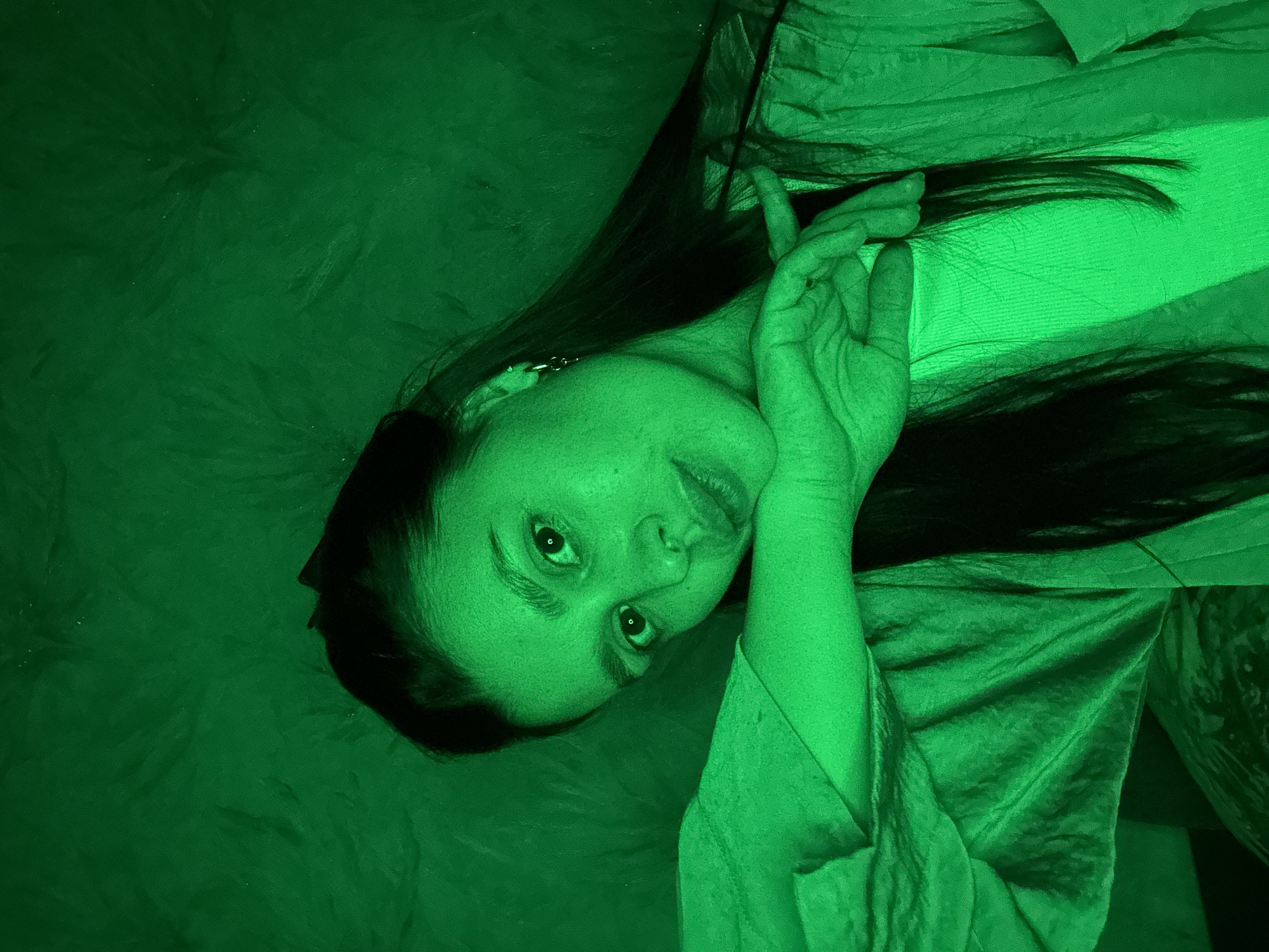 A woman with long dark hair is lying on a green surface, possibly a bed or cushion, with her head tilted towards the camera. She is wearing what appears to be a light-colored top under a draped, green fabric. Her hand is raised near her face, and she is looking directly at the camera. The scene is dominated by shades of green, creating a monochromatic effect. There is no visible evidence of the location being Chaikovsky, Russia.