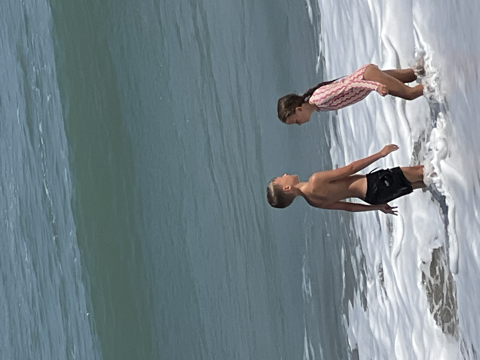 Two children, a boy and a girl, stand in shallow ocean water with foamy waves around their legs. The boy, on the left, is wearing black swim trunks. The girl, on the right, is wearing a pink and white striped long-sleeved swimsuit. They are facing each other and appear to be interacting. The ocean behind them has large, blue-green waves. The image appears to have been taken on a sunny day.
