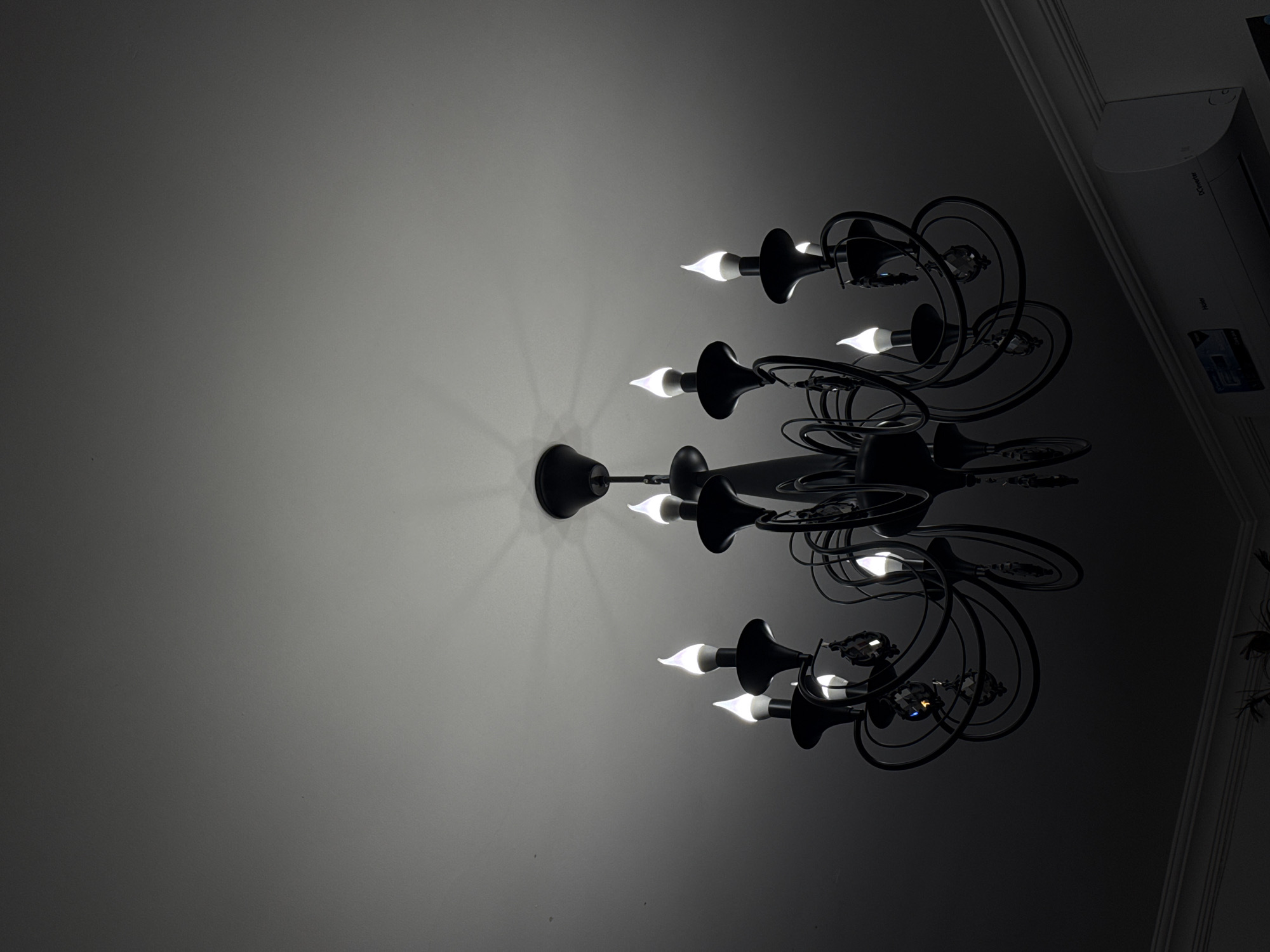 An ornate, black chandelier dominates the central and right portions of the image, viewed from below against a light grey ceiling. The chandelier features multiple gracefully curving arms, each ending in a candle-flame shaped light bulb that emits a bright, white glow. Several of the arms are adorned with small, faceted crystal-like decorative elements that reflect light. The bright bulbs cast distinct radial light patterns and illumination onto the textured, light grey ceiling, which gradually darkens towards the left and top right corners of the frame.
In the upper right corner, where the ceiling meets a wall, a darker area reveals architectural molding, suggesting a decorative cornice or trim. Partially visible on this upper wall or ceiling boundary is a white electronic device, likely an air conditioning unit. Faint text is discernible on this device; "DC Inverter" is visible, and below it, "Haier" can be partially made out, though it is cut off and very dim.
The scene is illuminated solely by the chandelier, suggesting it is either nighttime or a dark interior space. The overall aesthetic is one of elegant artificial lighting within a room, captured with a high contrast between the bright light sources and the surrounding darker elements. No human figures or discernible activity are present. The composition focuses entirely on the chandelier and its effect on the immediate surroundings.