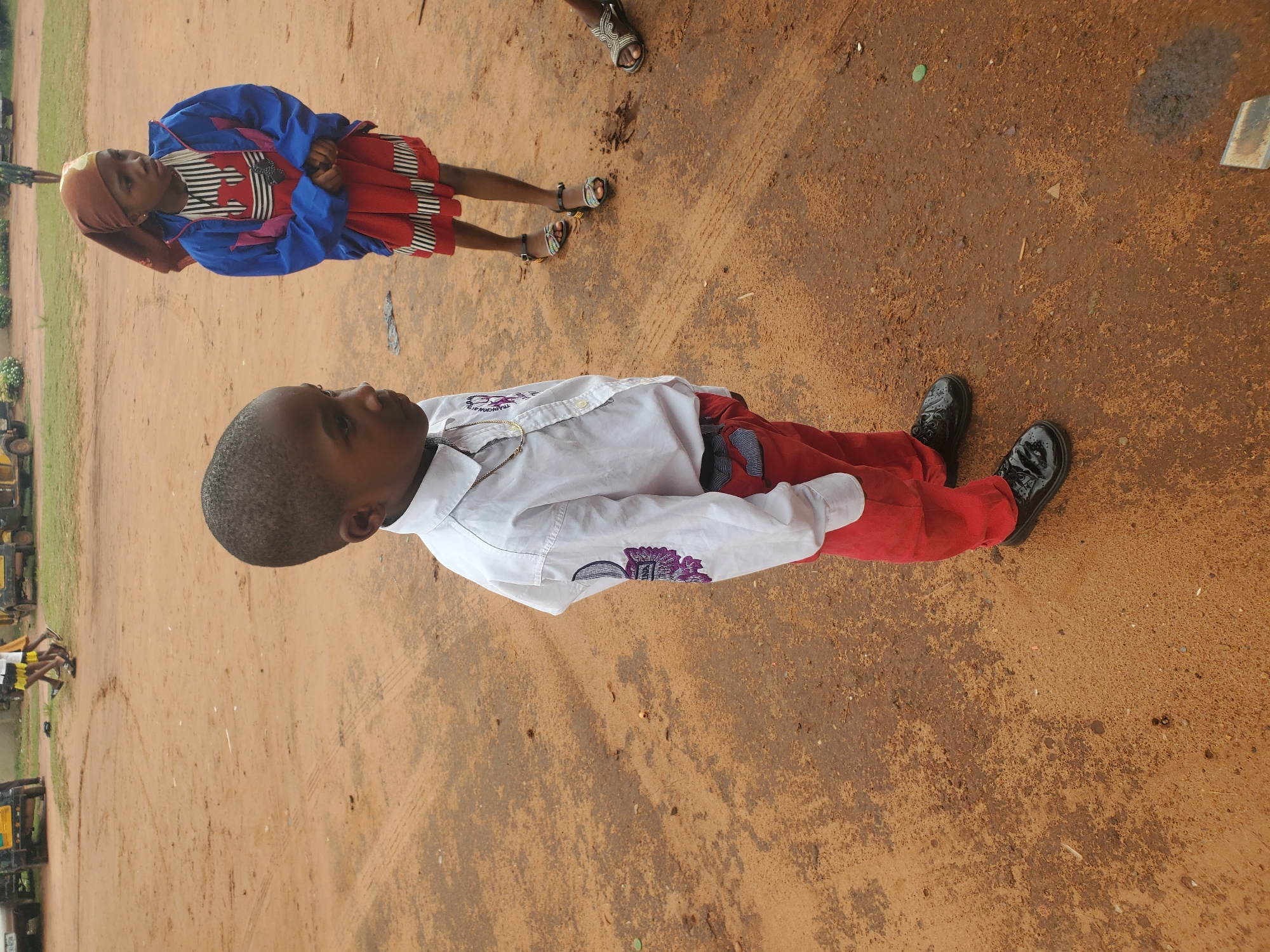 In Nsugpe, Nigeria, a young boy in a white collared shirt and red pants stands on a dirt ground, his body facing right. He wears black patent leather shoes and a gold chain. To his right, a young girl in a red dress and blue jacket, with a brown headscarf, also faces right. Another child's legs wearing sandals are visible in the background. The environment appears to be an outdoor area with sand and dirt, possibly a street or open space. Several yellow and black vehicles are parked in the background.
