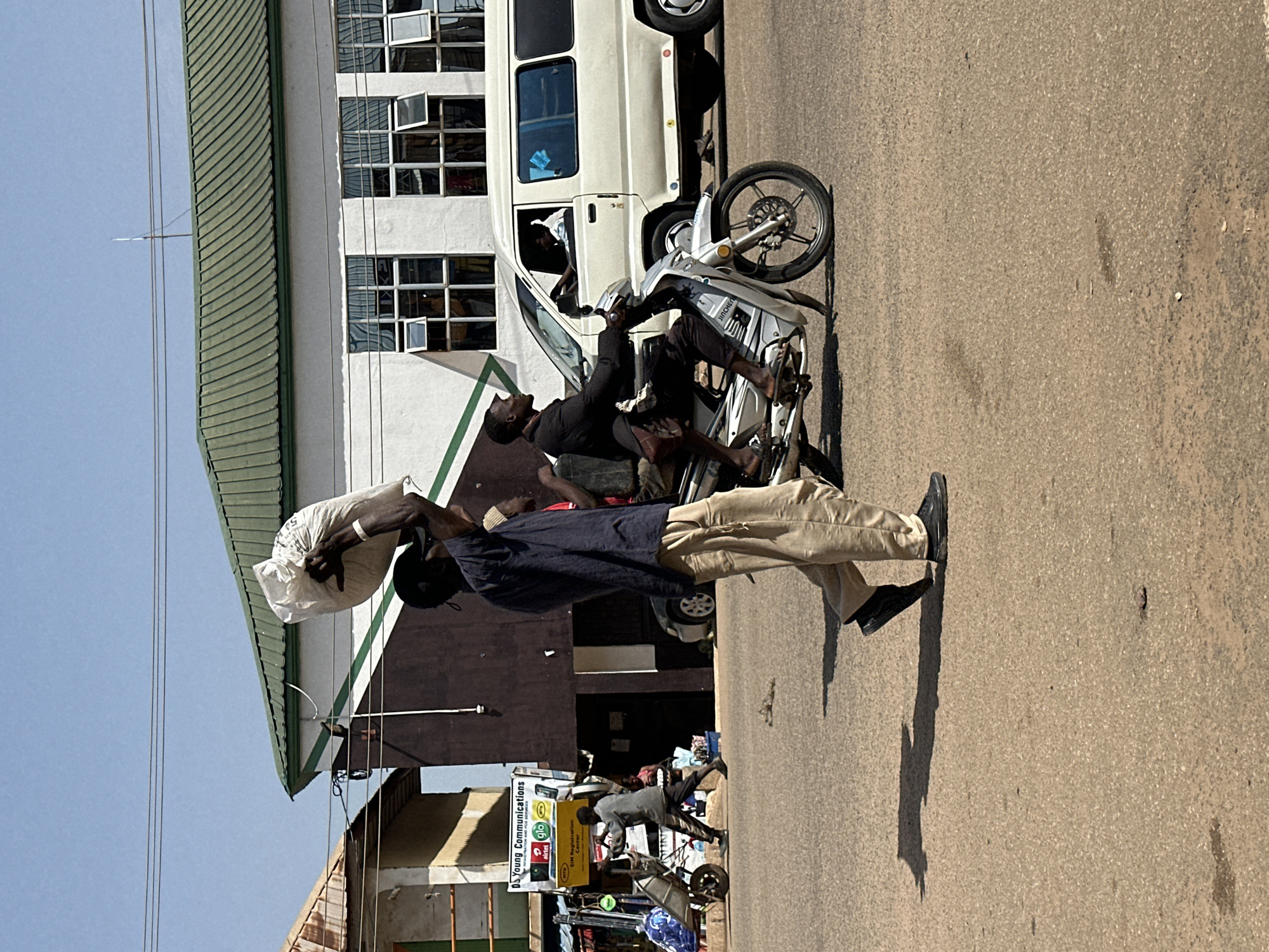 In Bukuru, Nigeria, a man walks down a street, carrying a large white sack on his head. He wears a dark blue short-sleeved shirt and light tan pants. To his right, a motorcycle carries two men; the driver wears a dark jacket and pants, and the passenger behind him is mostly obscured. A white van is parked on the right side of the road. In the background, buildings with green roofs line the street, and a yellow sign for "DS Young Communications" is visible. The sky is clear and blue. The street appears to be made of asphalt.