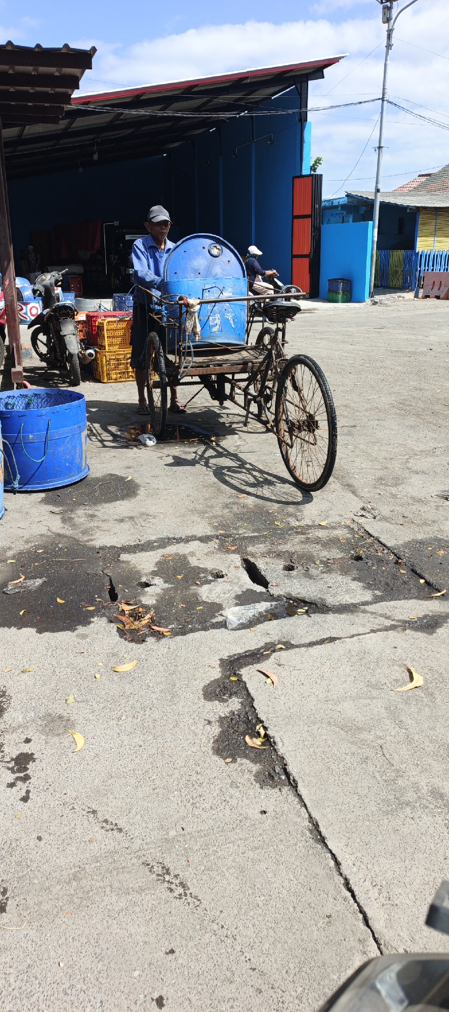 An elderly man wearing a blue shirt and a black cap is standing next to a blue barrel mounted on a three-wheeled cart. The cart appears to be made of metal and wood and has a rusty appearance. The man is holding onto the cart's handlebars. A second person, wearing a white cap, is seated on another three-wheeled cart behind the man, facing away from the camera. Several yellow and orange plastic crates are stacked near a motorcycle on the left side of the image. The scene takes place outdoors on a paved surface that shows signs of wear and tear, including cracks and water stains. In the background, there are buildings with blue and red facades, and a streetlamp. The sky is blue with scattered clouds. The image is a street scene in Puger, Indonesia.