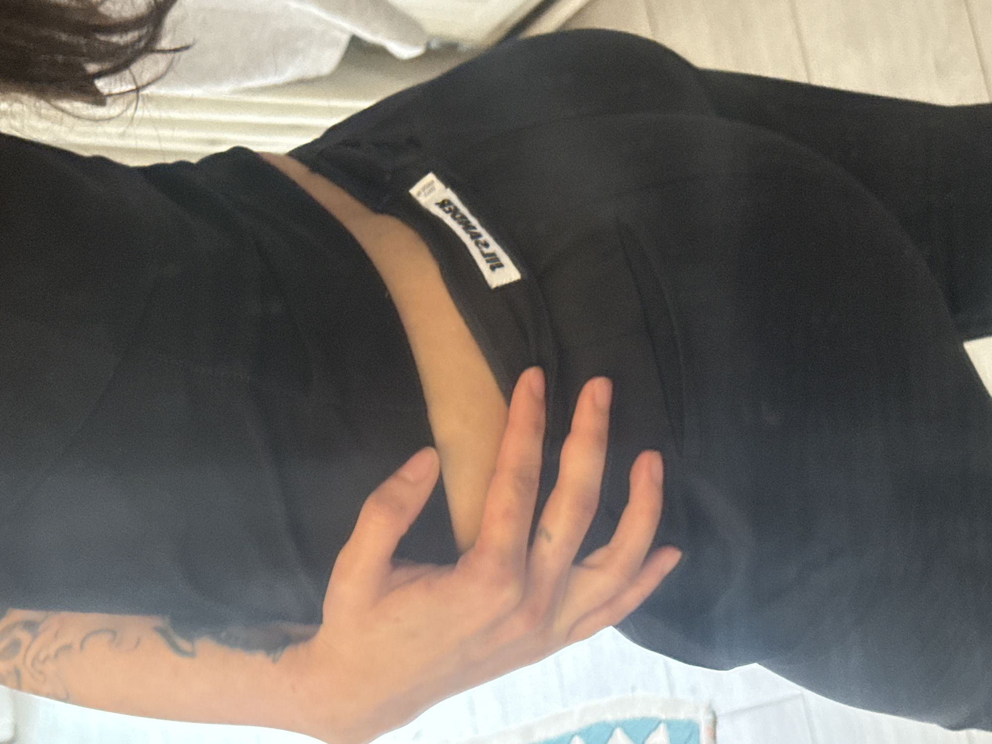 A person is wearing black pants with a white label on the back. The person's hand is on their hip, and a tattoo is visible on their arm. The location is not discernible from the image.