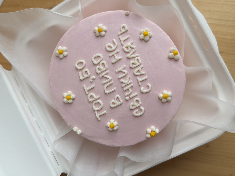 A circular cake with light pink frosting is centered within an open, white, rectangular cardboard take-out box. The cake's surface is decorated with white frosting text in Cyrillic script, reading "ТОРТ, БО В ПИВО СВІЧКИ НЕ СТАВЛЯТЬ", which translates to "CAKE, BECAUSE CANDLES AREN'T PUT IN BEER." Eleven small, white, five-petaled daisy-shaped decorations with yellow centers are distributed across the cake. Crinkled white tissue paper surrounds the cake inside the box. The box lid is partially open towards the top left. The entire assembly rests on a light brown wooden surface, possibly a table or countertop. No people, specific actions, or interactions are visible. The image provides no specific visual indicators for the city of Tel-Aviv, Israel.