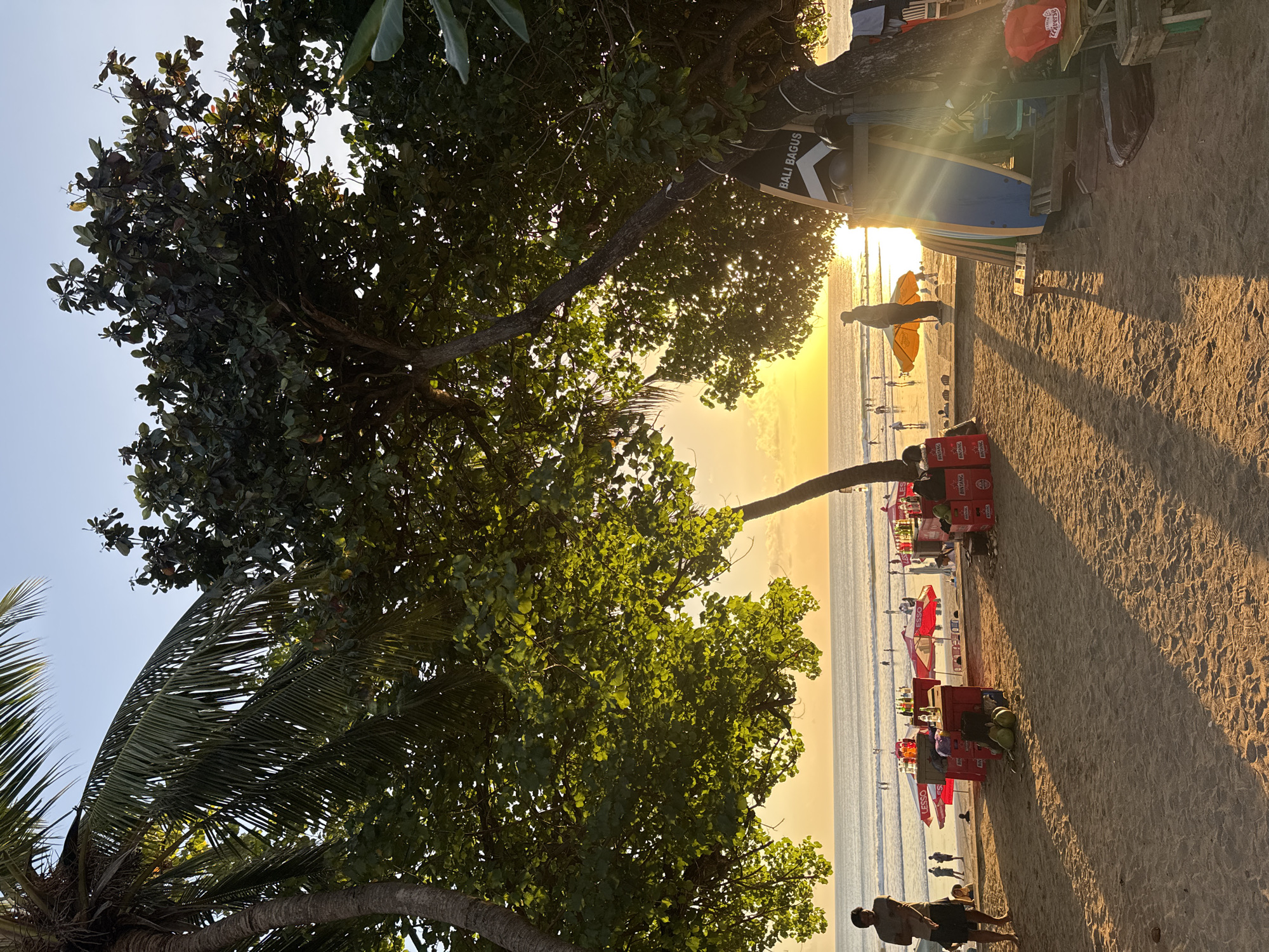 The image depicts a beach scene in Kuta, Bali, Indonesia during sunset. The foreground is dominated by sand with long shadows cast by trees. Several individuals are present on the beach, some standing and others seated. In the midground, there are beach umbrellas, stalls with drinks and coconuts, and surfboards leaning against a structure. People are visible walking along the shoreline and in the water. The background features the ocean with gentle waves and a vibrant sunset sky. The overall atmosphere is relaxed and leisurely.